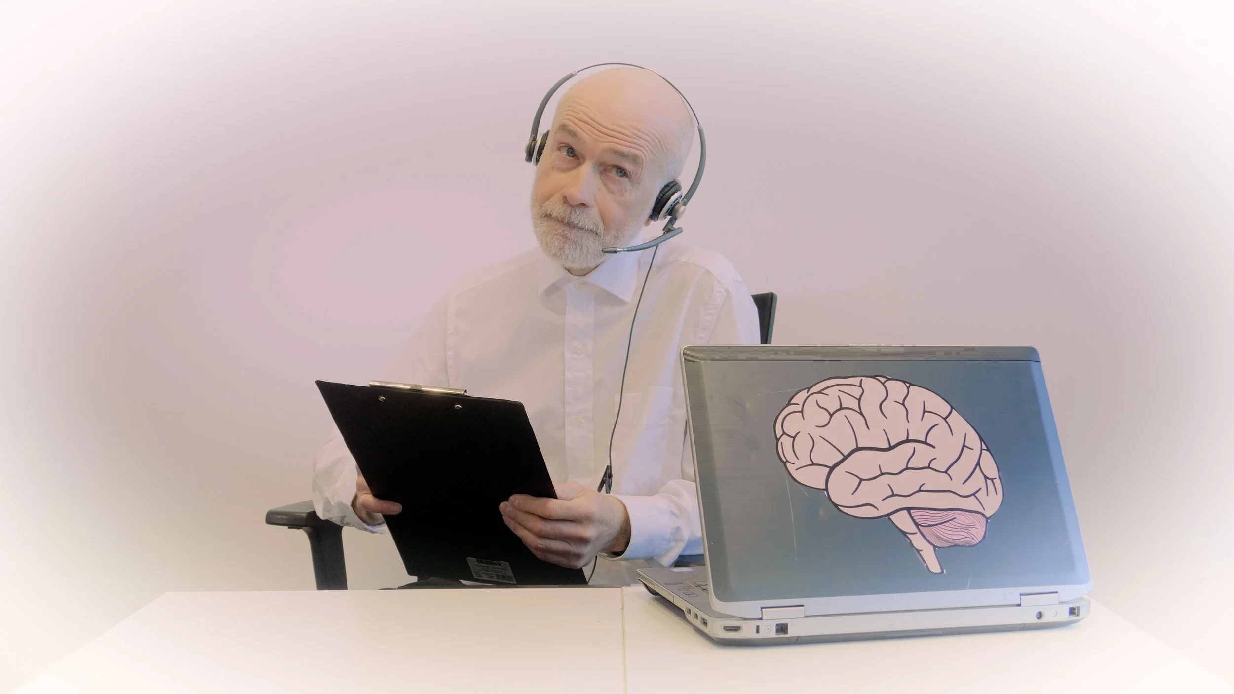 An elderly man with a bald head and white beard, wearing a white shirt, is sitting at a desk with a laptop that has a brain illustration on the lid. He is holding a tablet and wearing a headset with a microphone, looking slightly puzzled or curious.