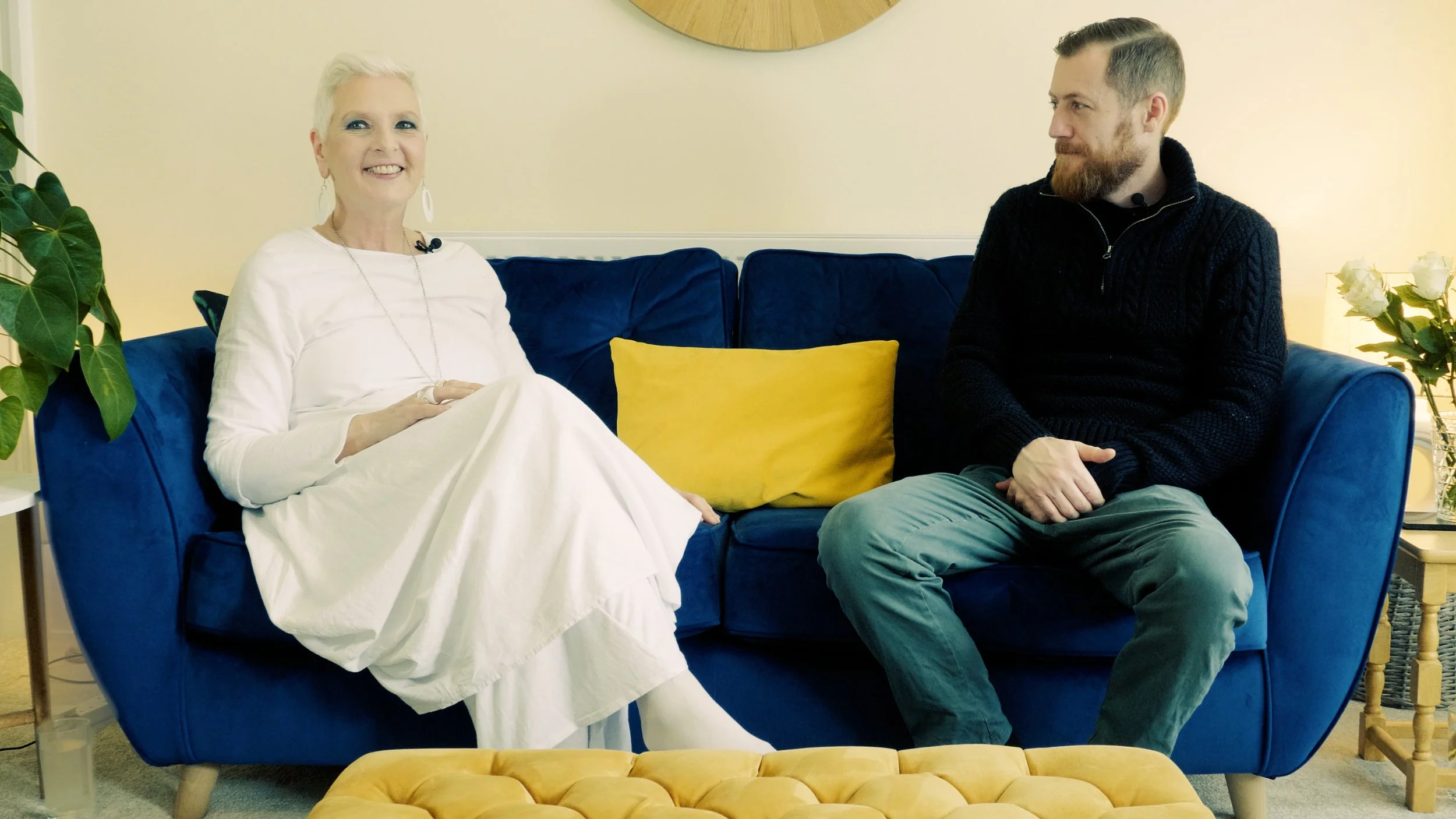 A woman in a white dress sits on a blue couch with a hellow cushion next to a man with a beard, a blue jumper and blue jeans