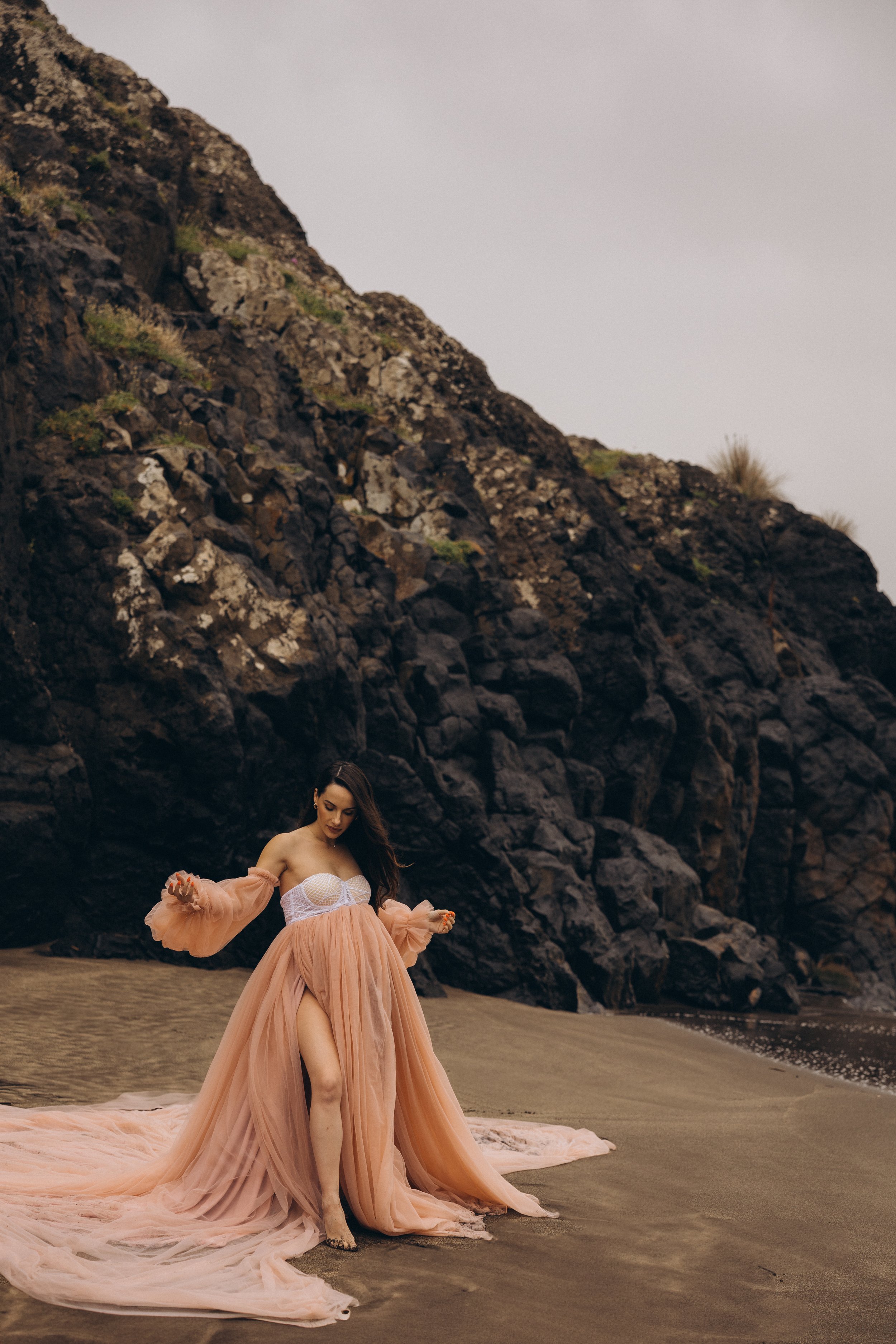 Woman in a flowing peach gown posing on a sandy beach beneath rugged dark volcanic cliffs