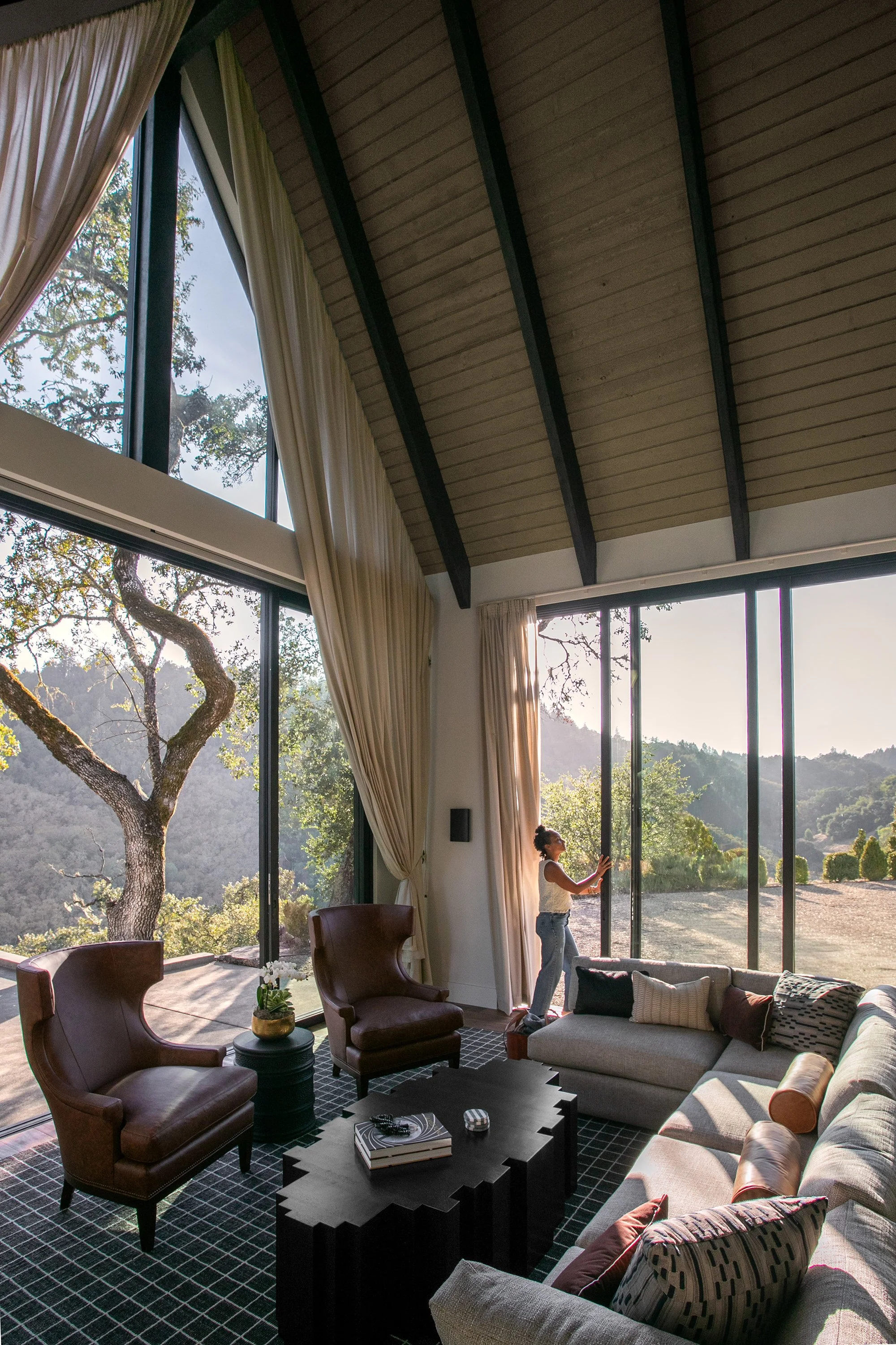 Women is opening a glass wall panel system in a room with draperies with views of oaks and surrounding hills.