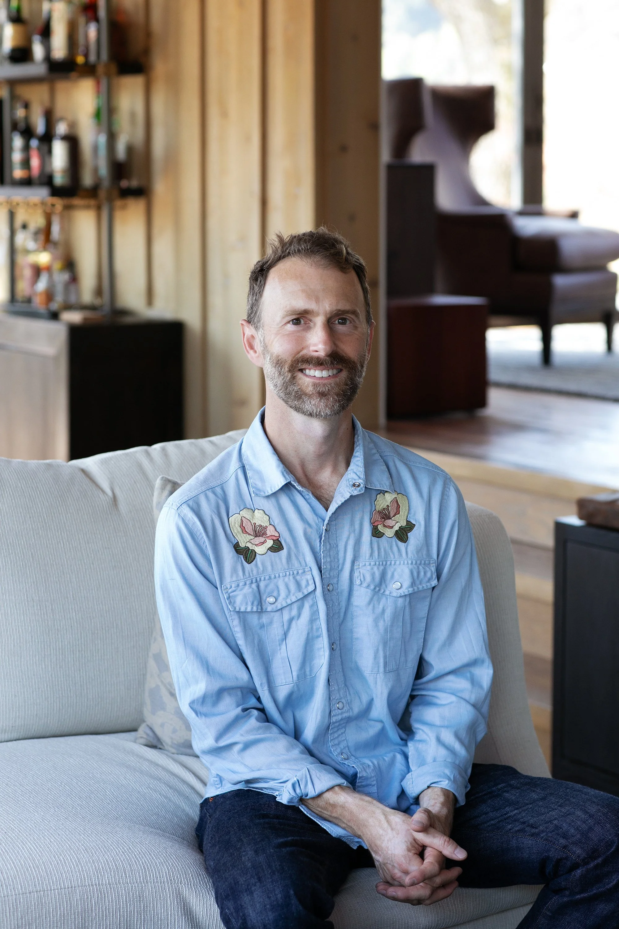 A man sitting on a cream-colored couch in a cozy, wooden interior with a bar shelf and a large window behind him, smiling at the camera.