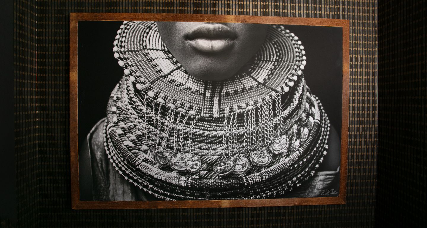 Framed black-and-white photograph of a Samburu woman wearing an elaborate layered beaded collar with dangling chains and pendants, displayed against dark wallpaper with a narrow vertical striped pattern.
