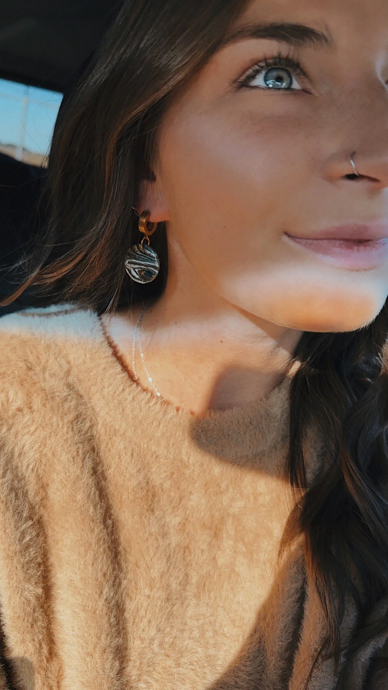 A close-up of a woman with blue eyes, dark brown hair, wearing earrings with striped circular pendants, a silver nose ring, a delicate necklace, and a tan fuzzy sweater, with sunlight illuminating her face.