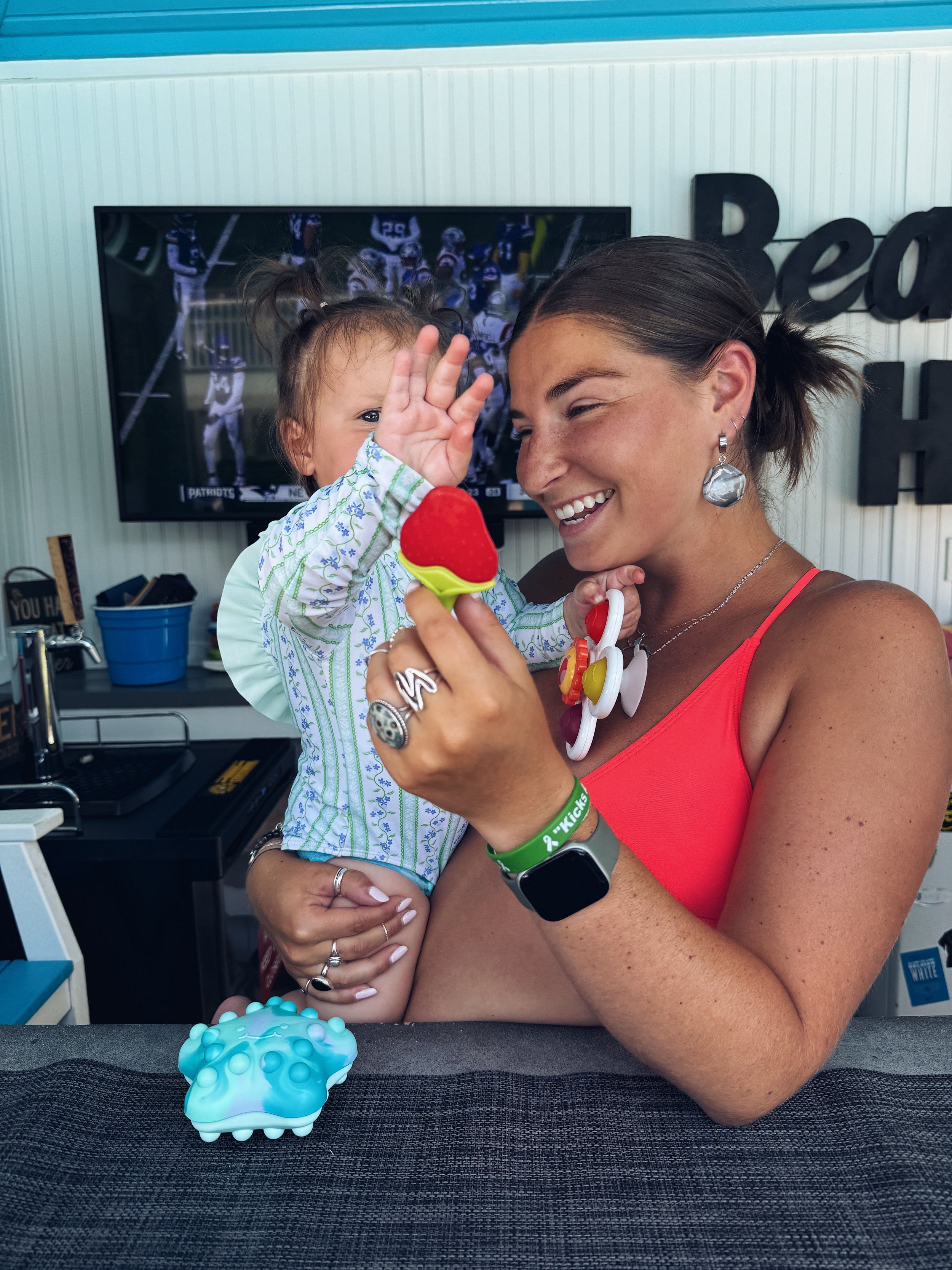 Woman holding a young girl in a colorful indoor space, with a TV screen and decorative signs in the background, and a blue textured object on the table in front.