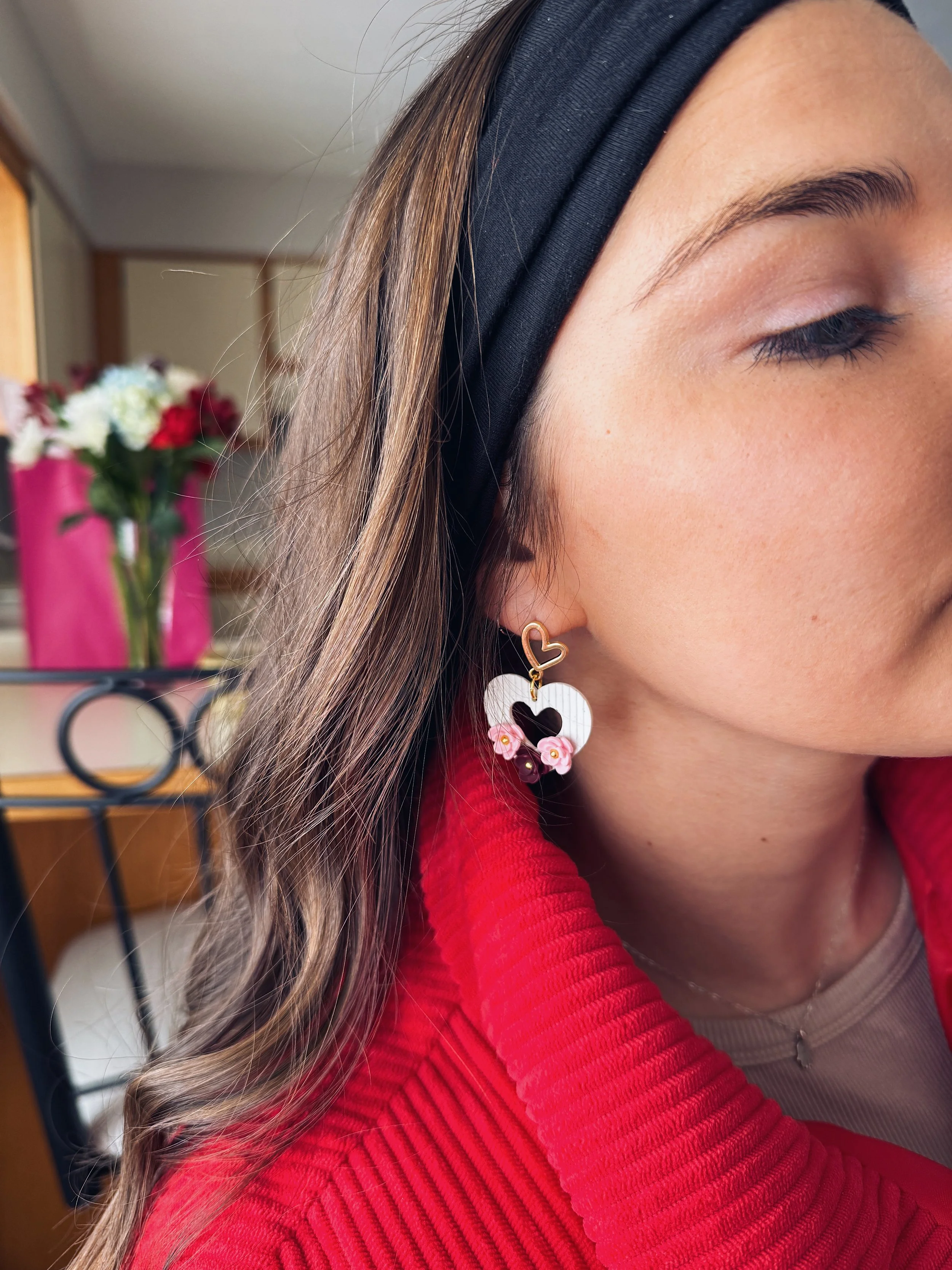 Close-up of a woman's face showing her eye, with a black headband, wearing a heart-shaped earring with pink flower decorations, red clothing, and a pearl necklace, with a blurred background featuring flowers in a pink bag.