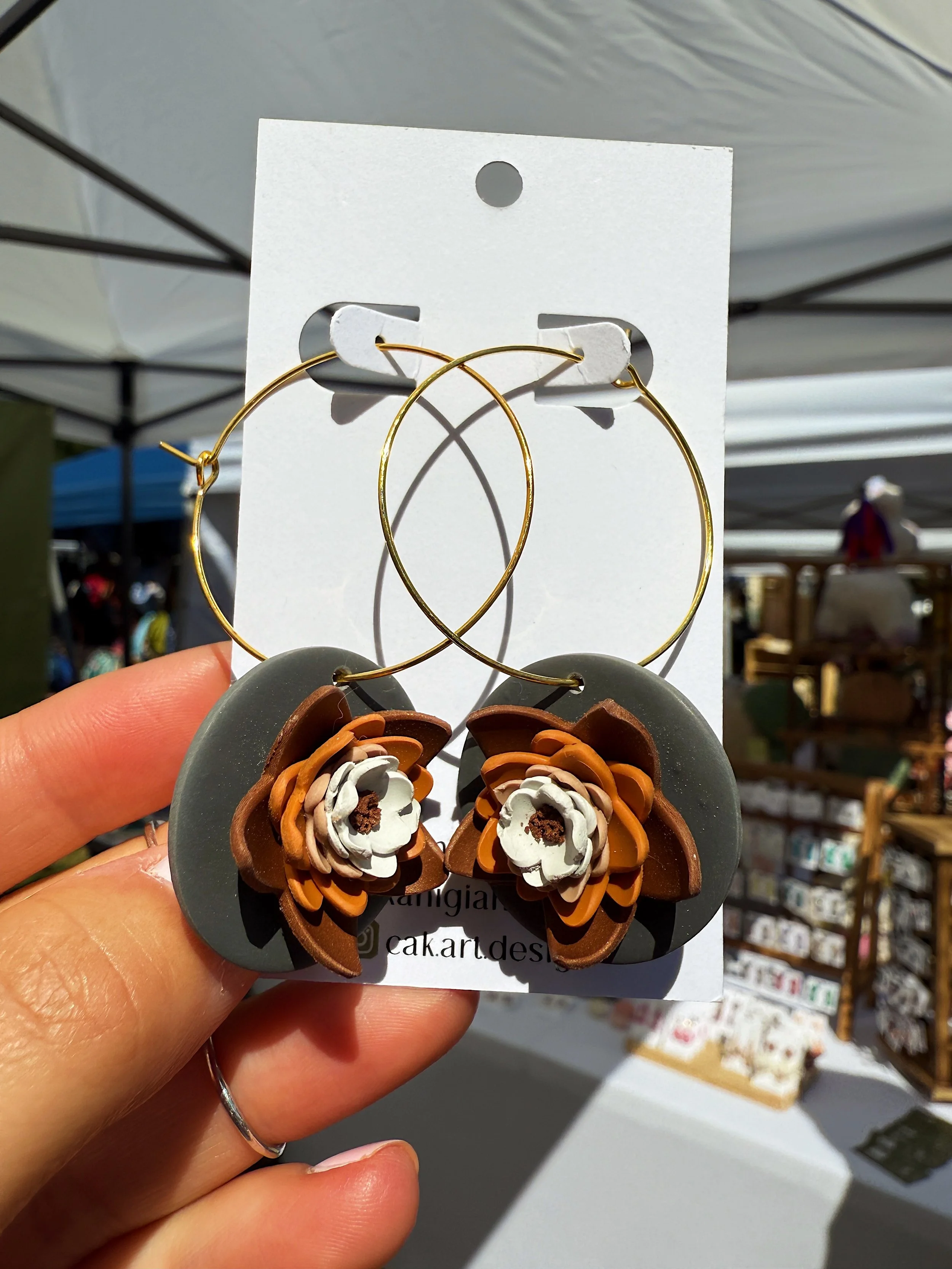Hand holding a display card with a pair of large circular earrings, featuring floral designs made of layered leather in brown, white, and beige, with gold hoops at the top.