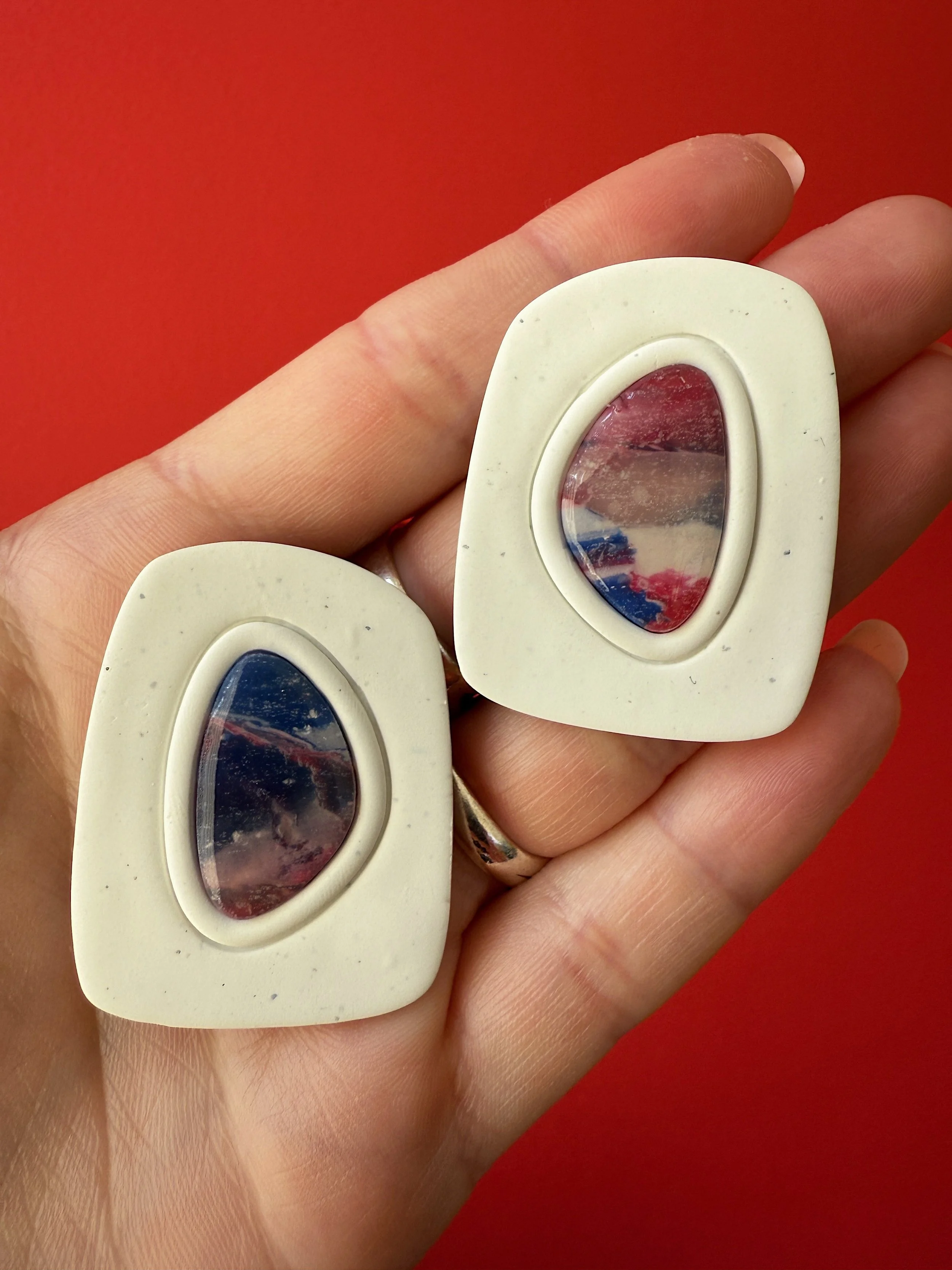 A hand with a silver ring holding two large rectangular earrings with oval-shaped, reflective surfaces, set against a red background.