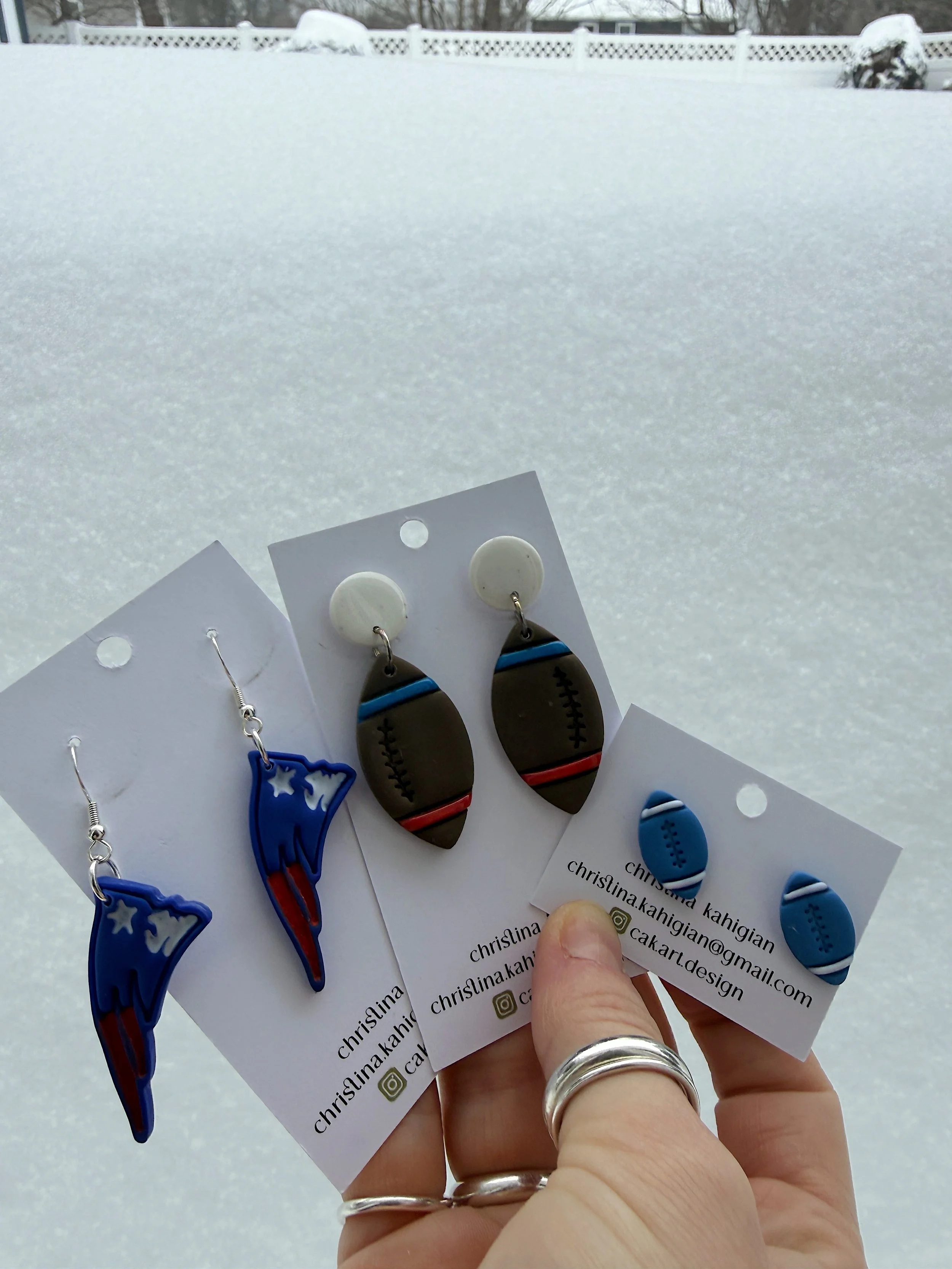 Three pairs of handmade earrings with different sports-themed designs, held in front of a snowy backyard with a white fence.