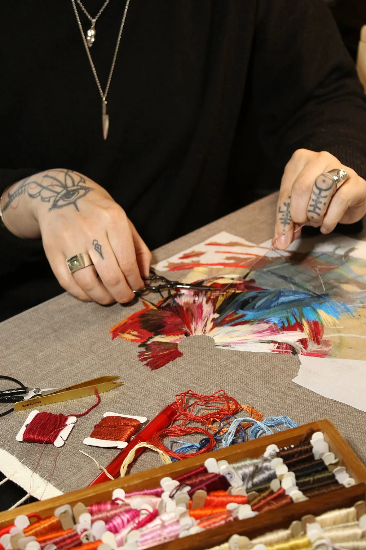 Lucy Newman  working on a colorful embroidery or fabric art piece, surrounded by embroidery threads and tools on a table.