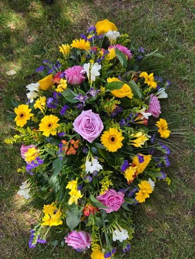 A colorful arranged floral casket spray with pink, purple, yellow, and white flowers on green grass.