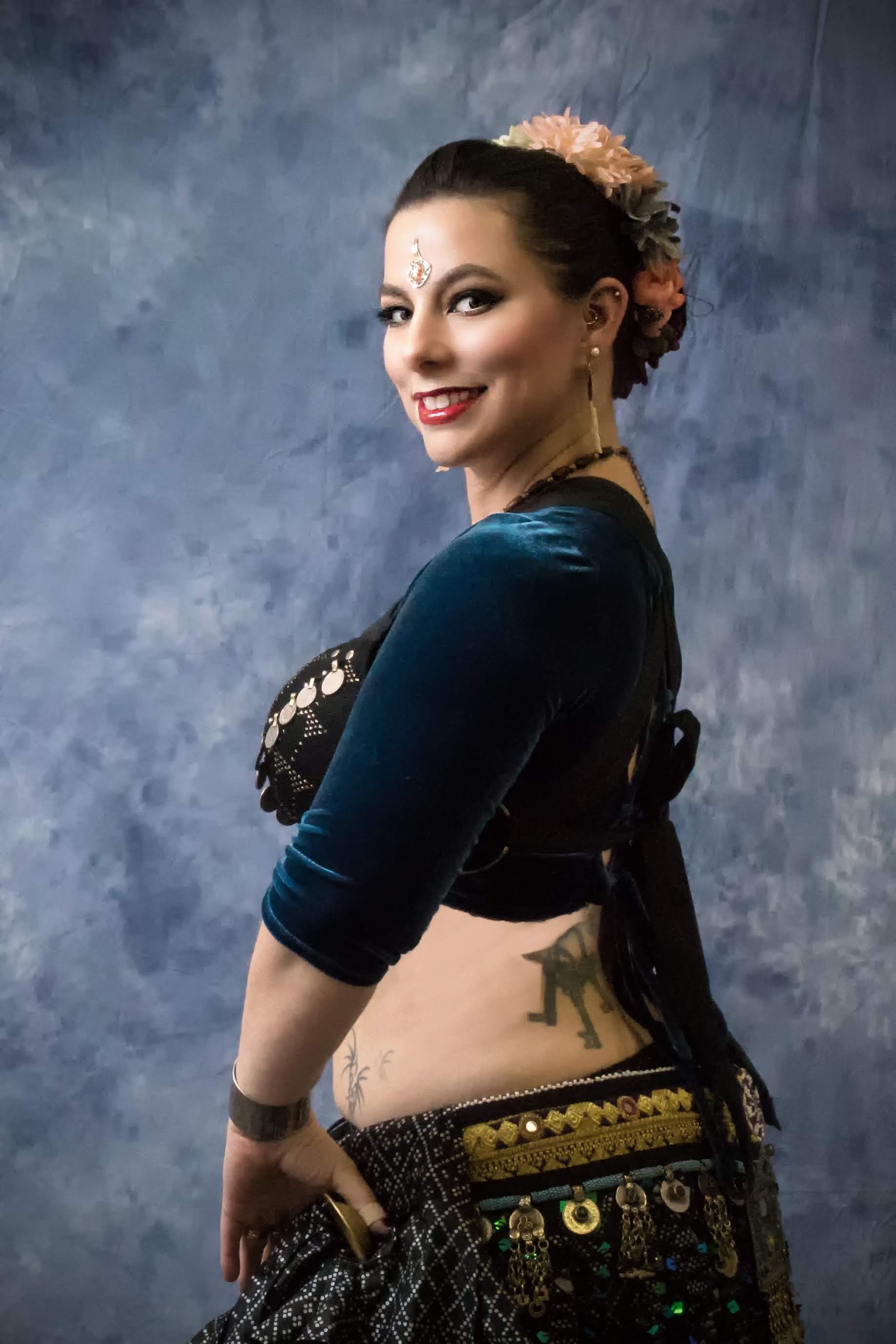 A woman dressed in traditional dance attire, with flowers in her hair, jewelry, and tattoos, posed against a textured blue background.