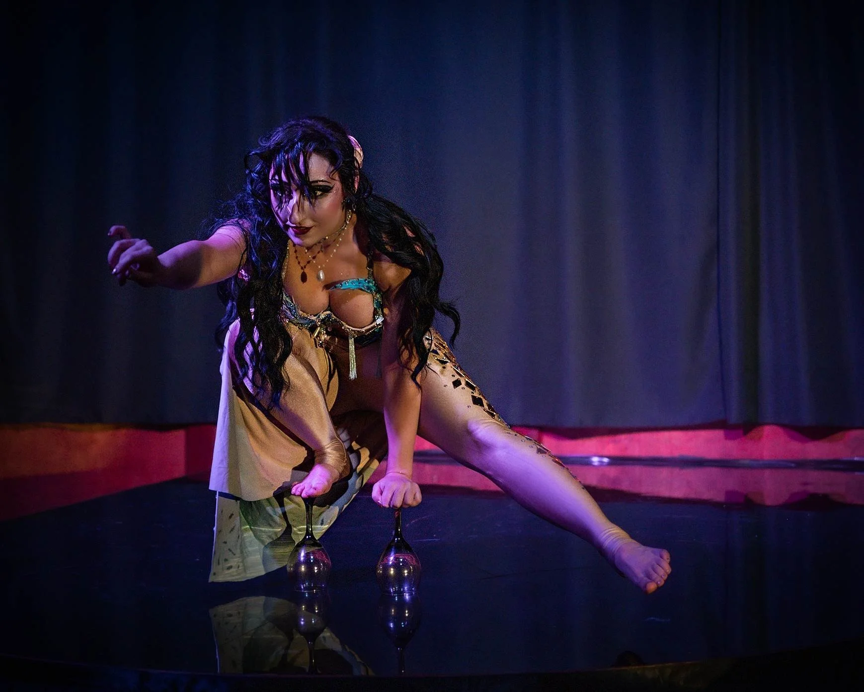 Performer with dark curly hair, decorated with flowers, wearing revealing costume, balancing on wine glasses, on stage with curtain background.