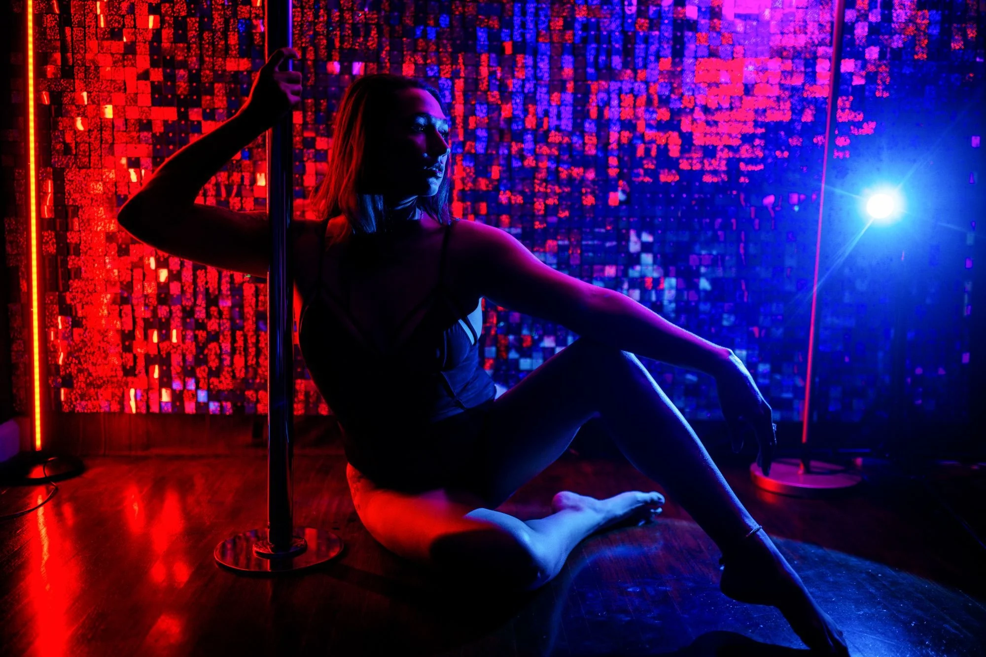 Woman seated beside a pole in a colorful red and blue neon boudoir set.