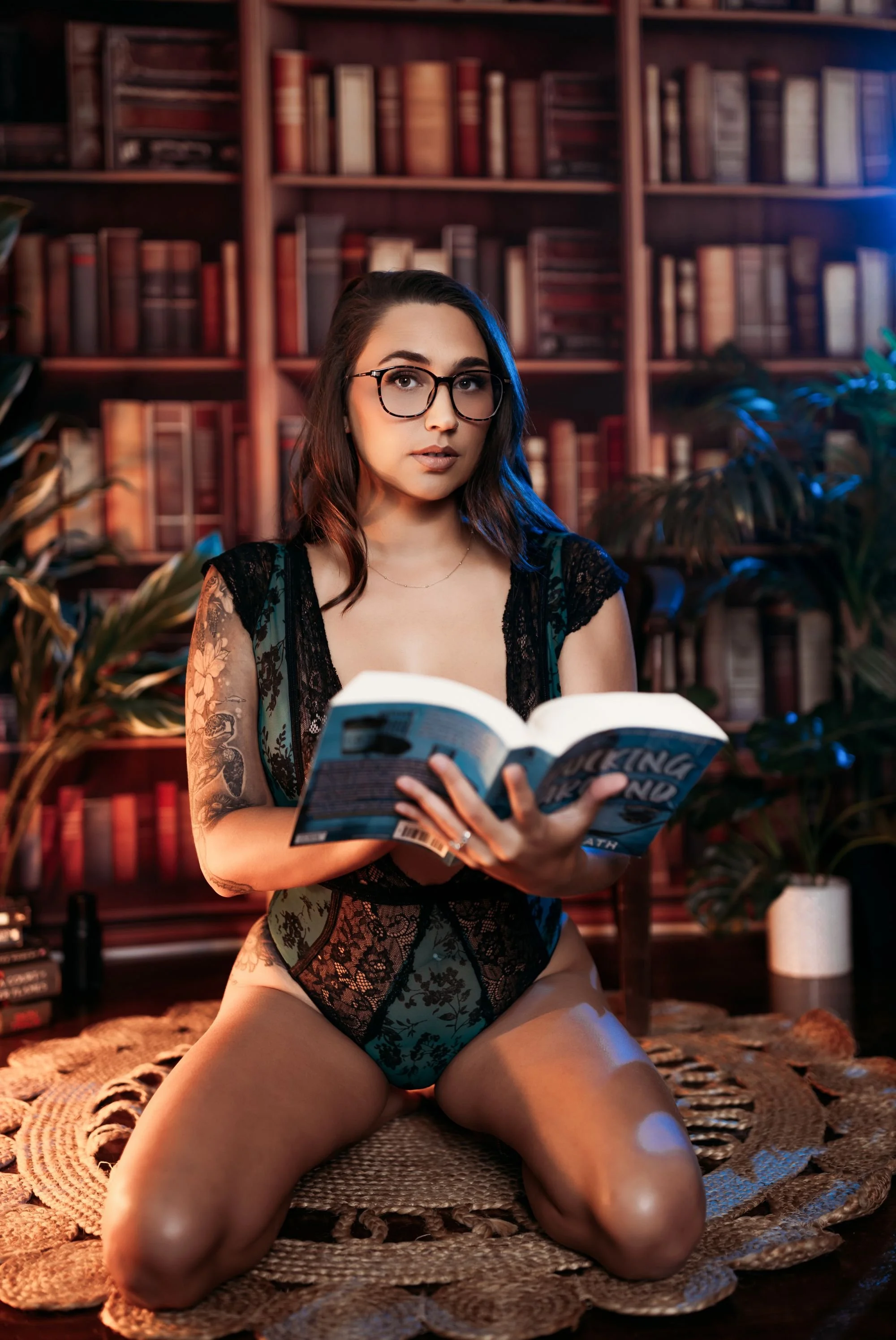 Woman wearing glasses and a lace bodysuit holding an open book in the studio’s moody library set.