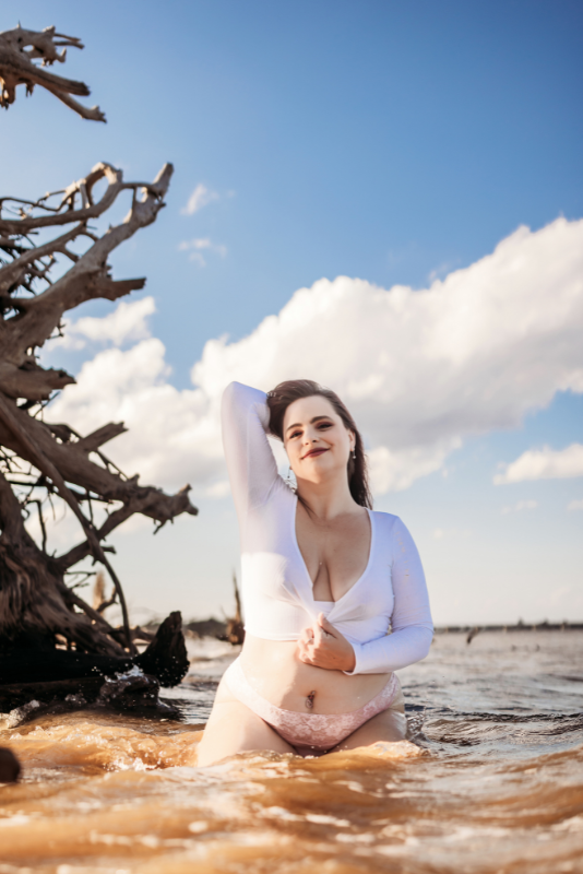 Woman in a white long-sleeve top sitting in shallow water by the beach.