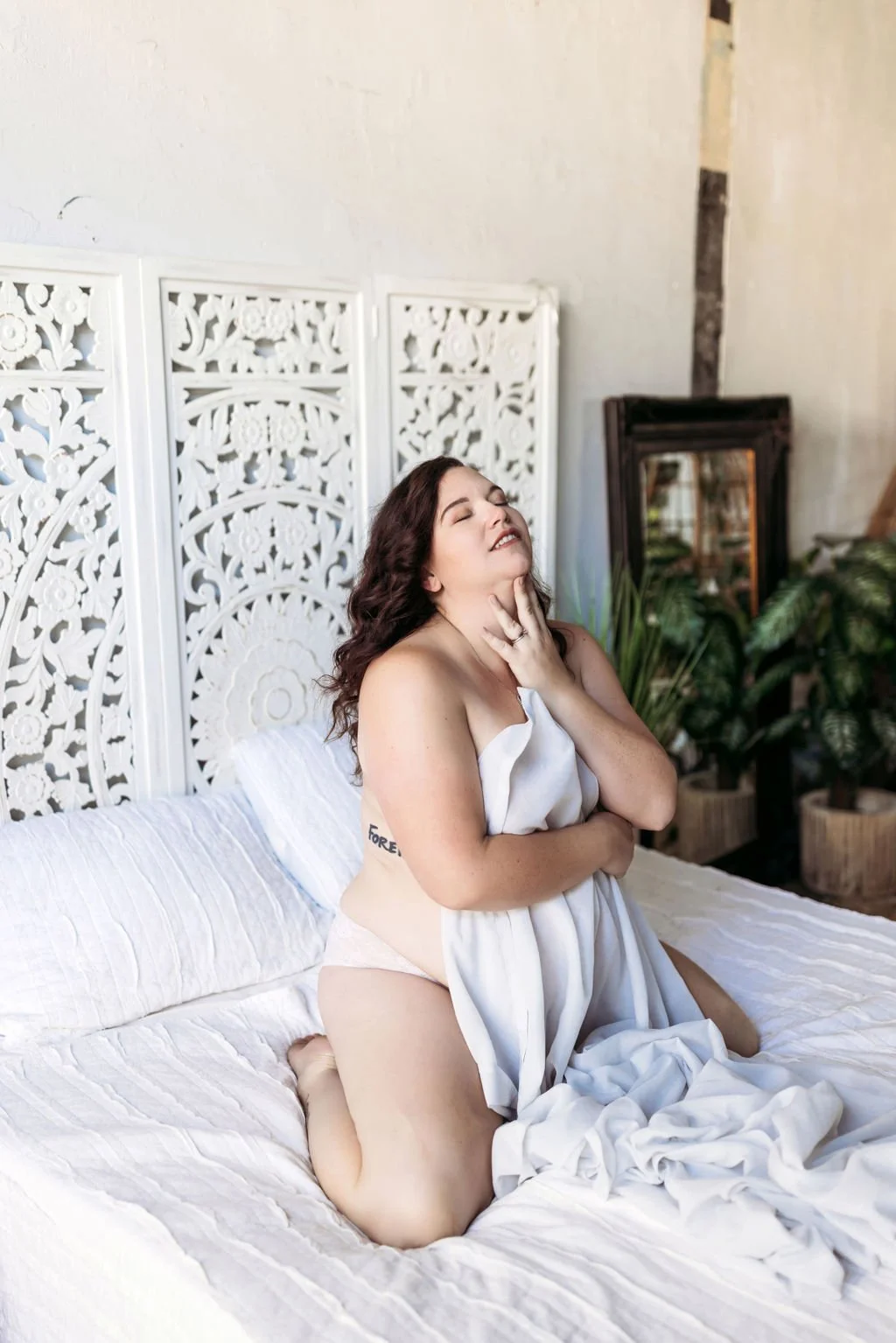 Woman kneeling on a bed while posing with a white sheet during her boudoir session
