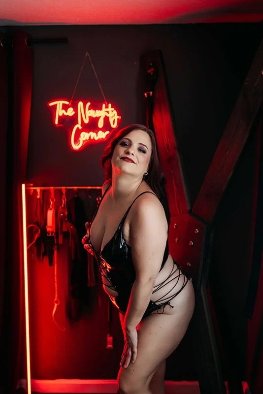 Woman wearing black leather lingerie posing beside a St. Andrew's cross in Hype Girl Boudoir's red neon set