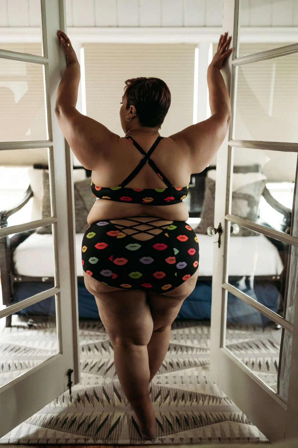 Woman in colorful kiss-print lingerie posing with her back to the camera between French doors