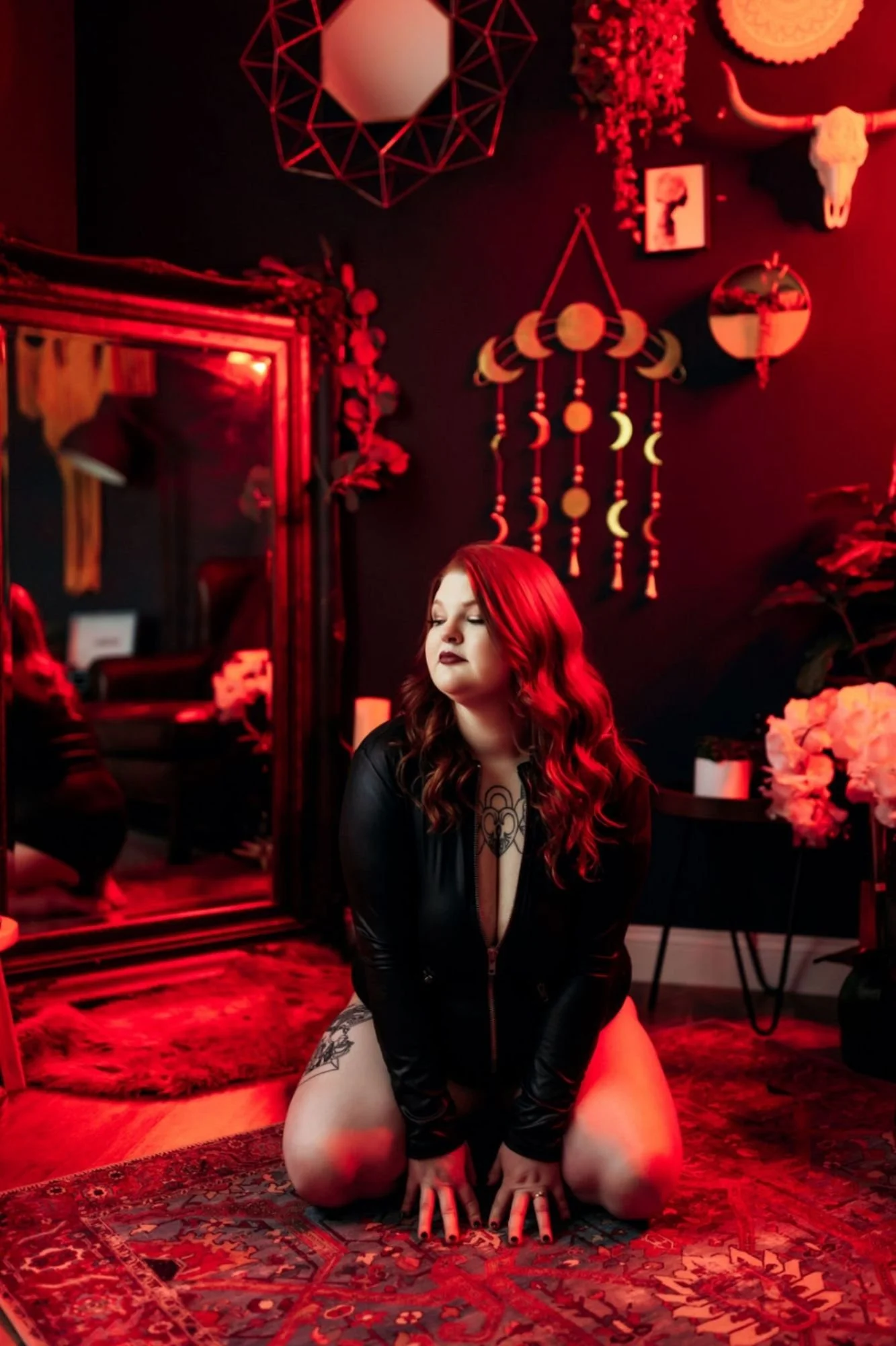 Red-haired woman kneeling on a patterned rug in a red-lit studio.