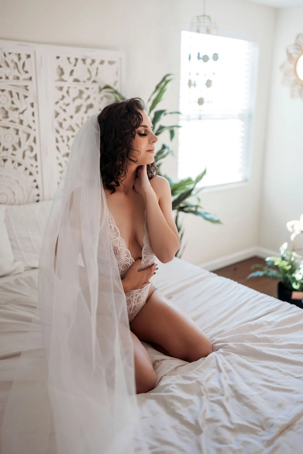 Woman in a white lace bodysuit and veil kneeling on a white bed.