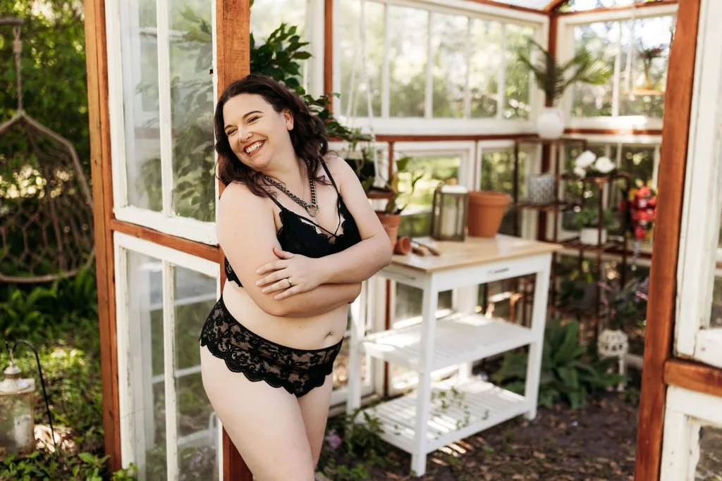 Woman laughing in black lingerie while posing inside the greenhouse during her boudoir session