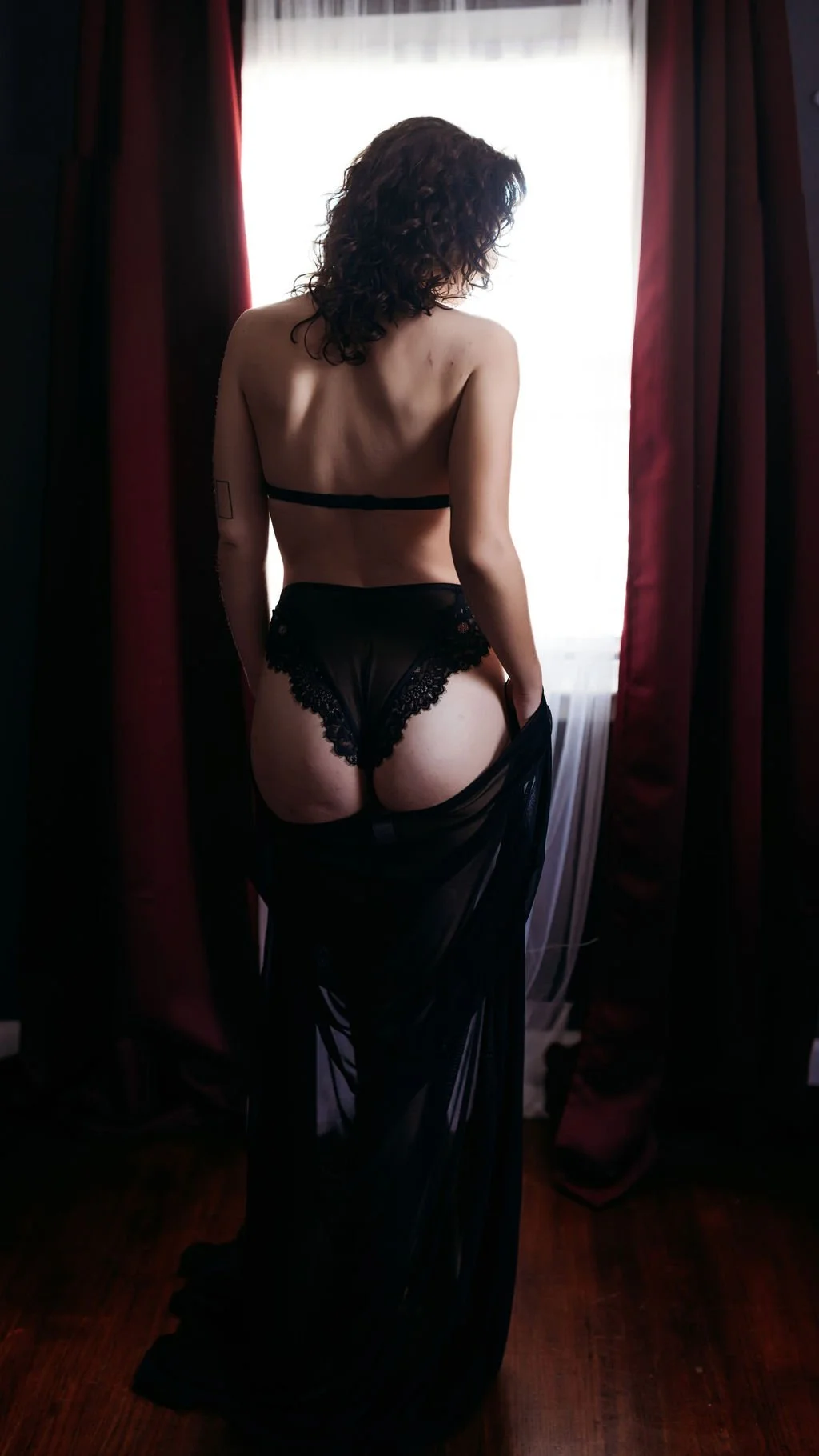 Woman in black lingerie standing with her back to the camera beside a bright window.