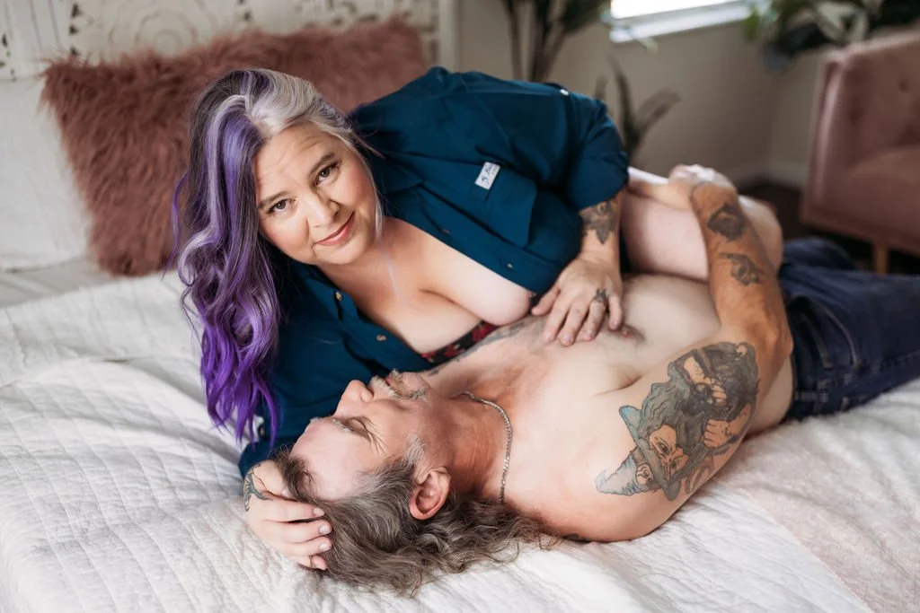 Older tattooed couple embracing while lying together on a bed during their boudoir session