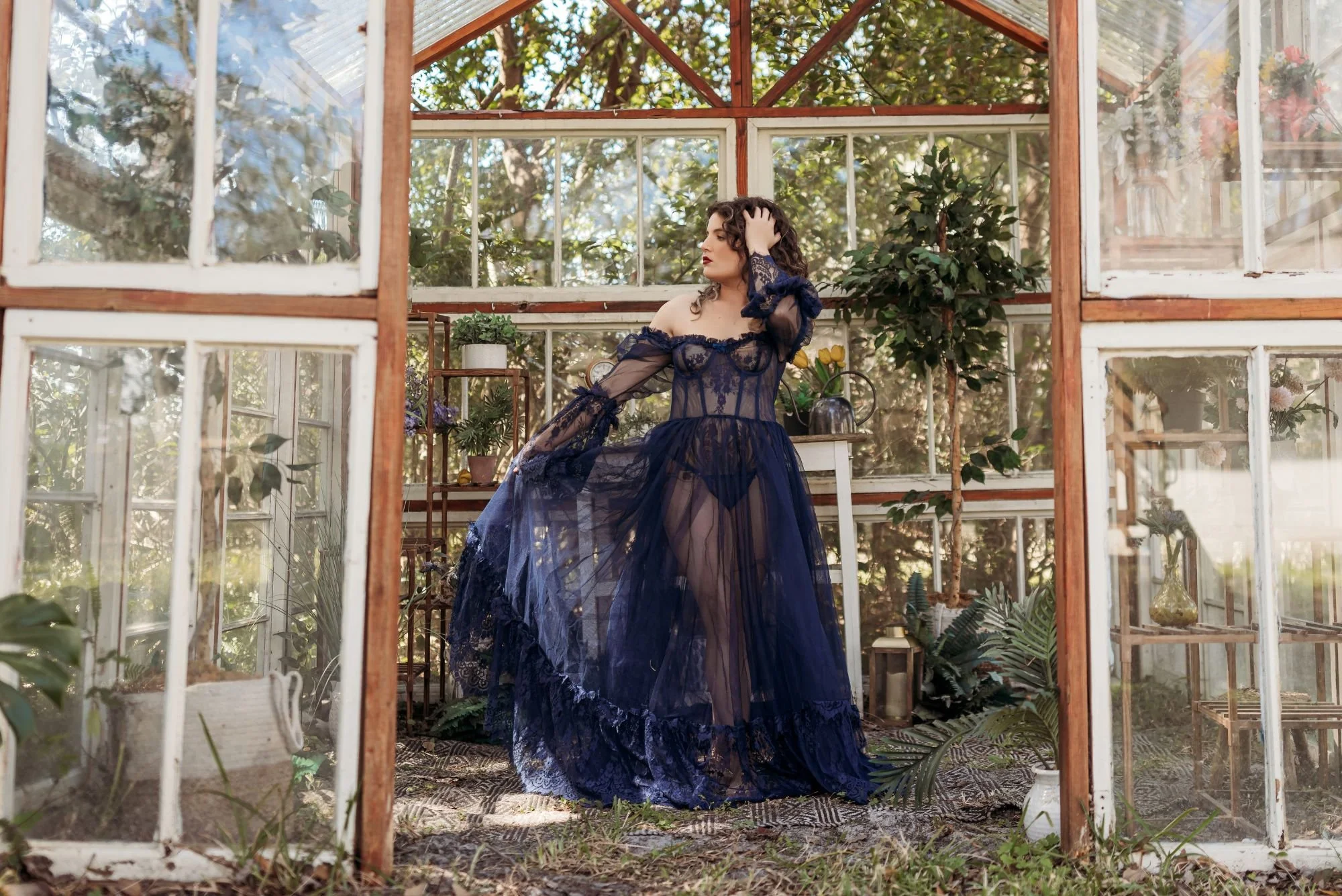Woman in a flowing navy sheer gown standing in a greenhouse.