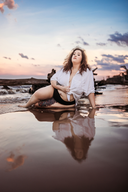 Woman in a white button-down shirt sitting in shallow water at sunset.
