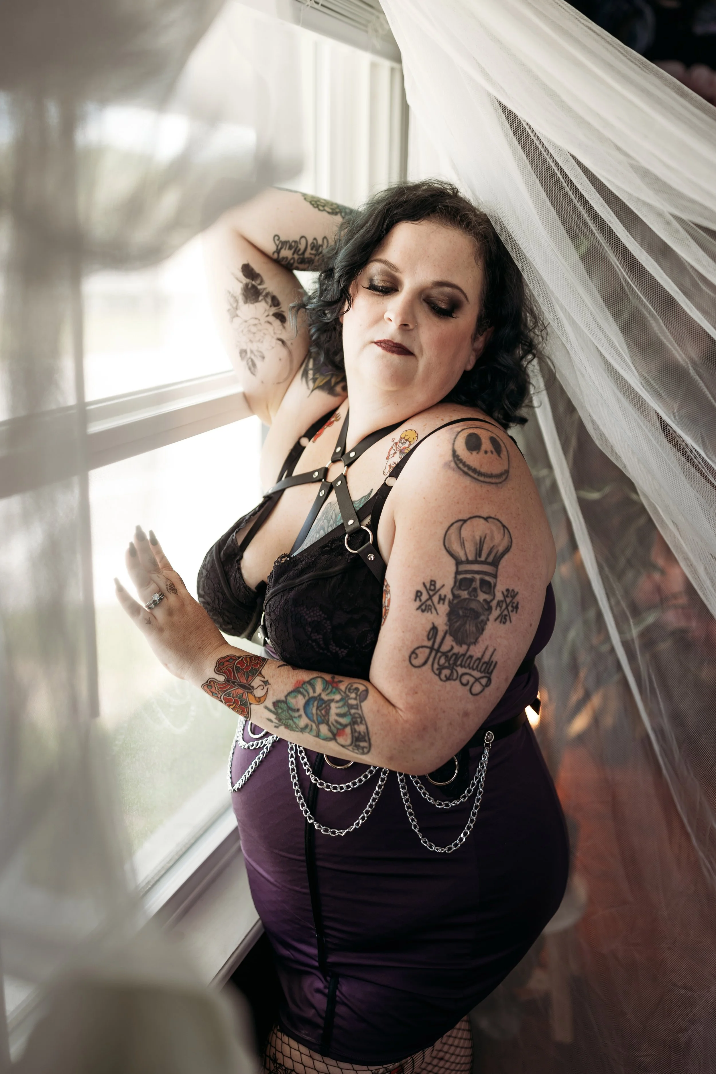 Tattooed woman in a dark outfit posing beside a bright window and sheer curtains.
