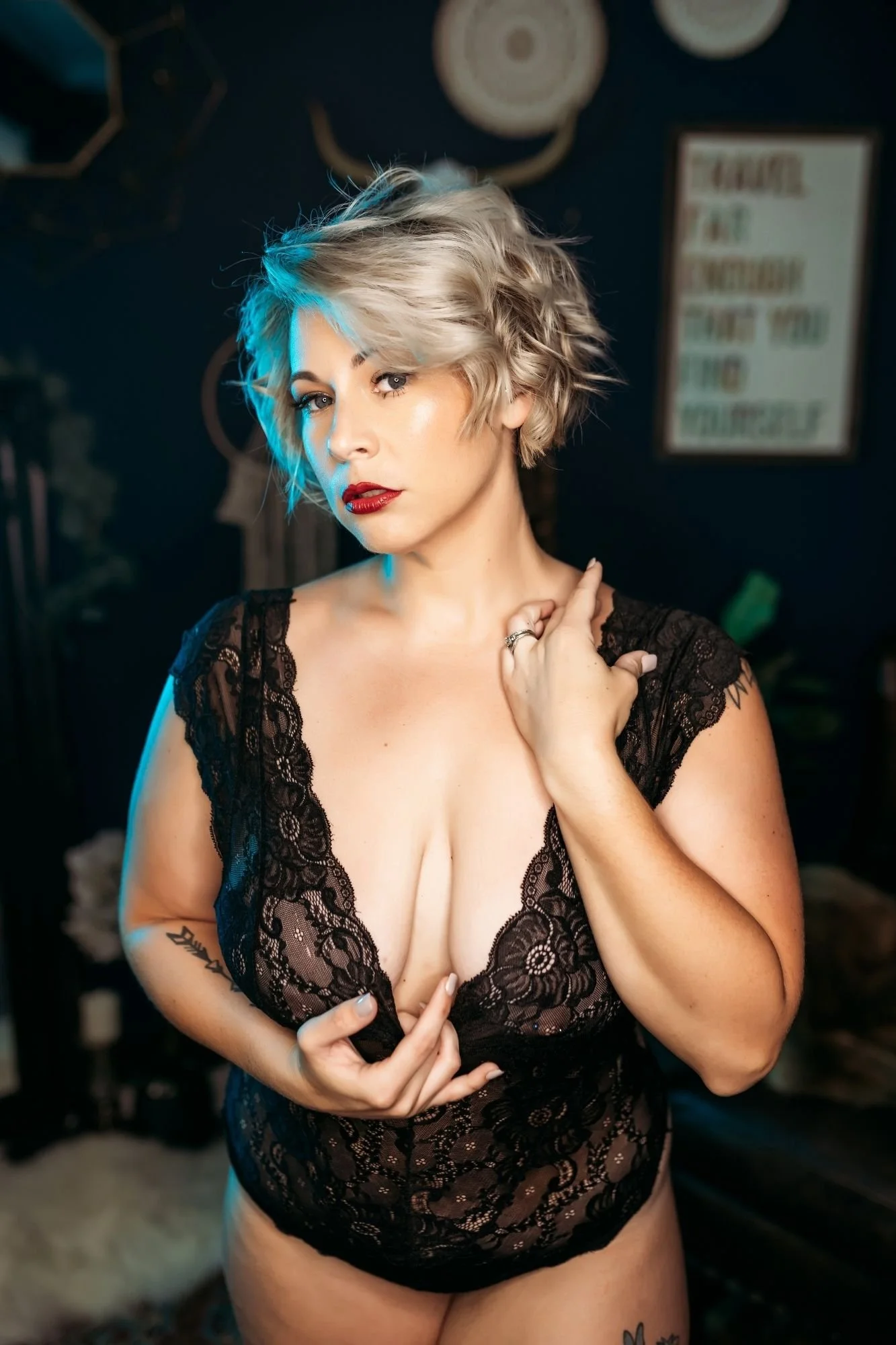 Woman with short blond hair posing in a black lace bodysuit against a dark background.