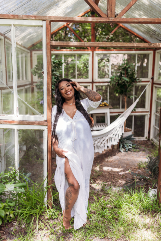 Woman in a flowing white dress standing beside a hammock in a greenhouse.