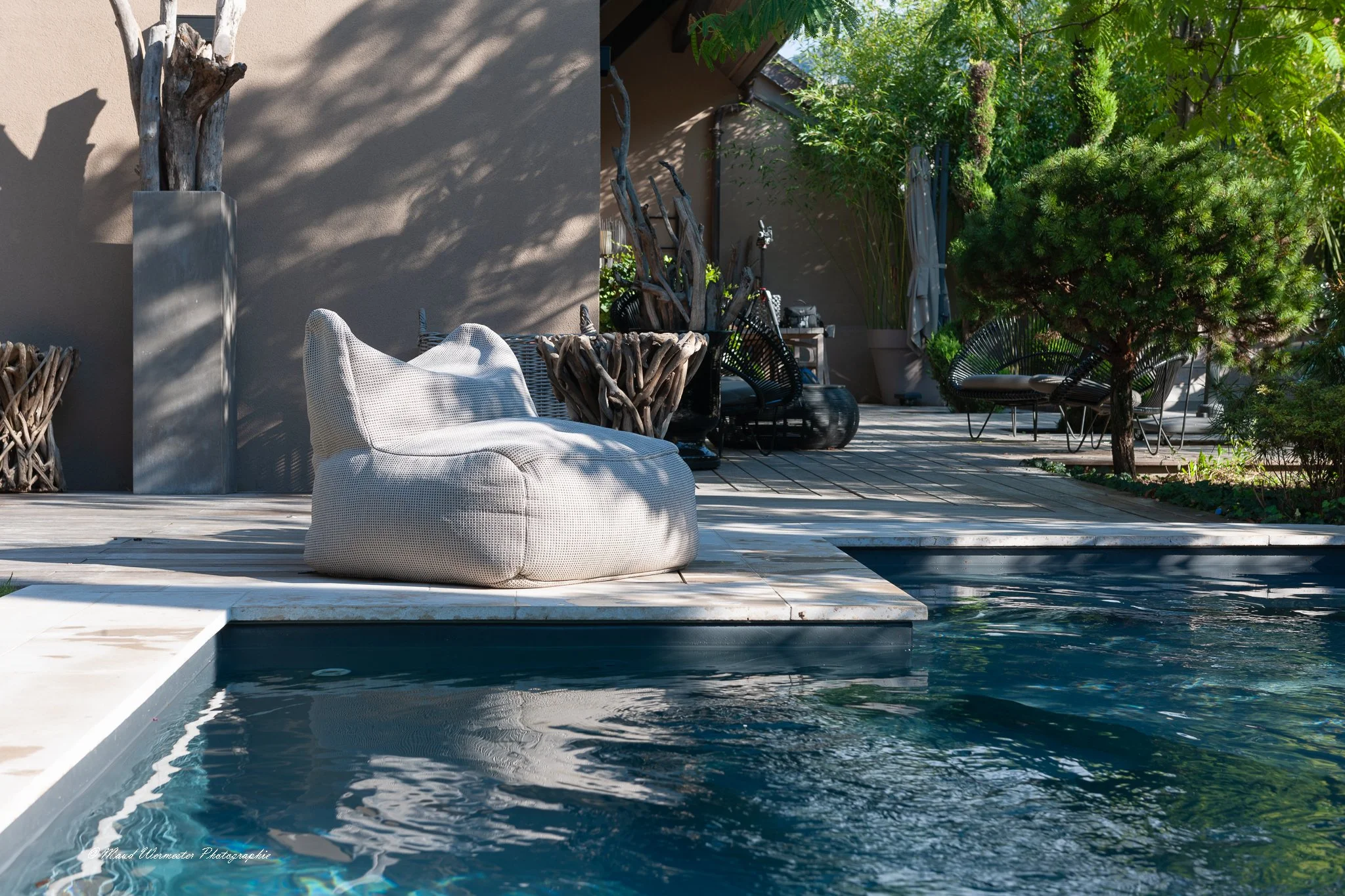 Une terrasse près d'une piscine avec un fauteuil en tissu blanche, des chaises en rotin noir, des plantations verdoyantes, et un mur en béton beige, dans un cadre extérieur ensoleillé.