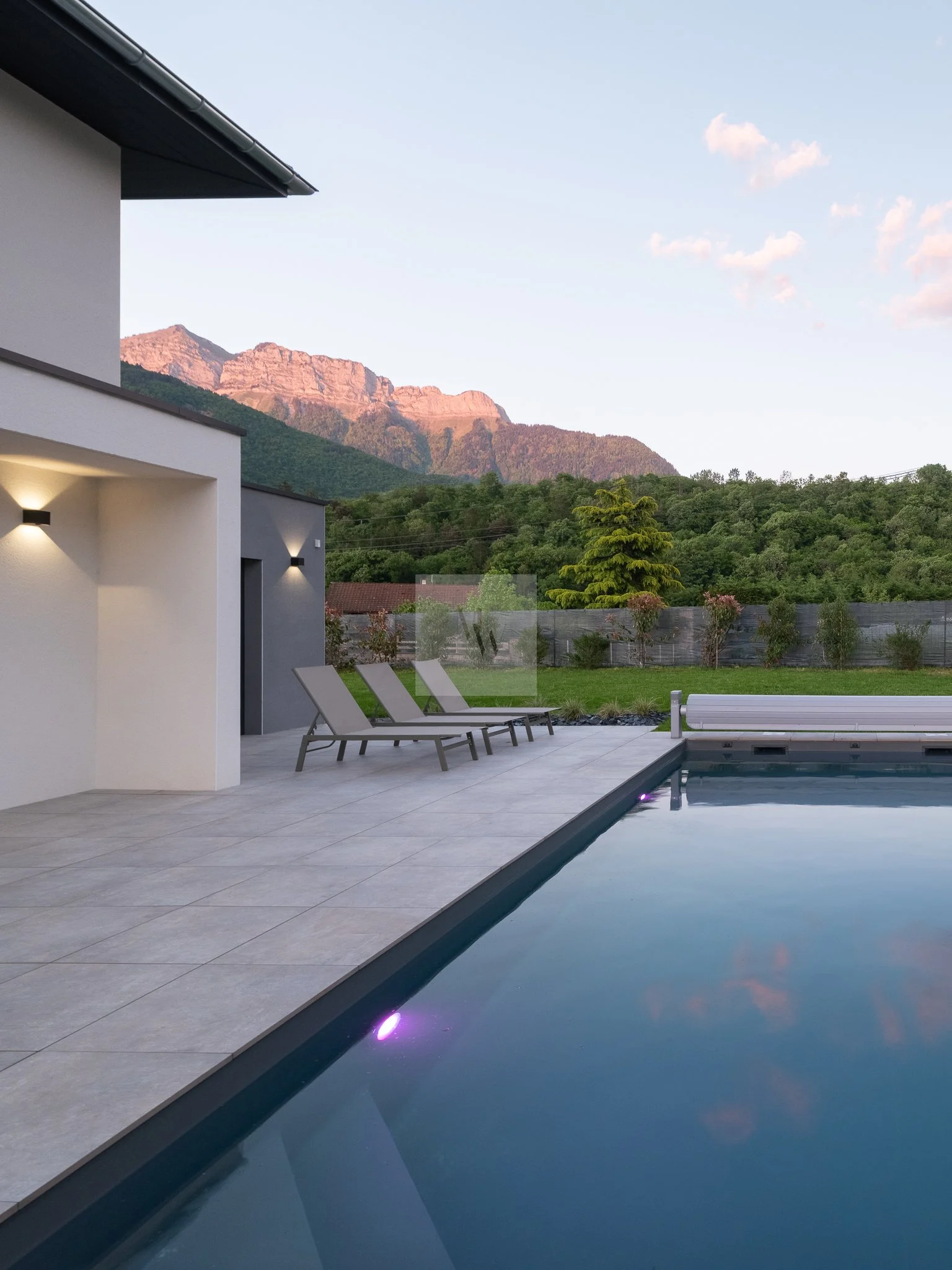 Piscine extérieure dans un jardin avec des chaises longues, maison moderne, montagnes au loin, ciel clair. Photographie immobilière premium pour locations saisonnières à Annecy