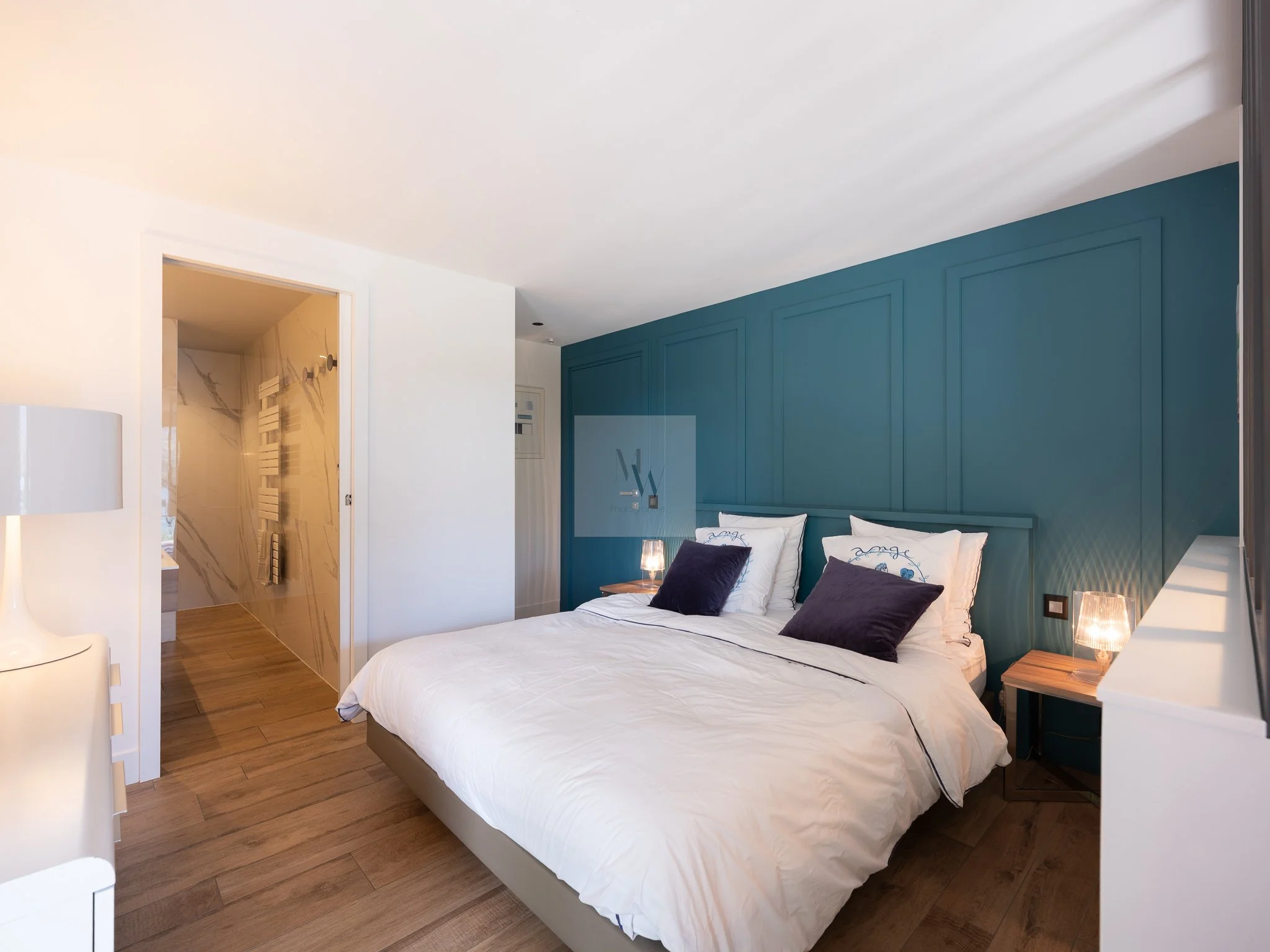 Chambre moderne avec lit double, murs bleus, lampes de lecture, et porte menant à une salle de bains en marbre blanc. Shooting photo professionnel pour biens immobiliers rives du lac d'Annecy