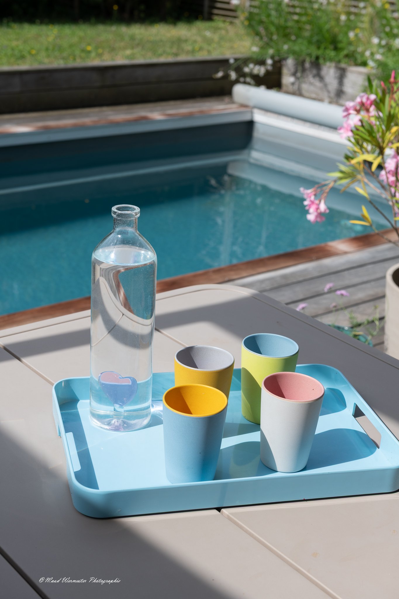 Verre d'eau transparent et plusieurs gobelets colorés (bleu, jaune, blanc, rose) sur un plateau bleu clair, placé sur une table extérieure. En arrière-plan, une piscine et des plantes