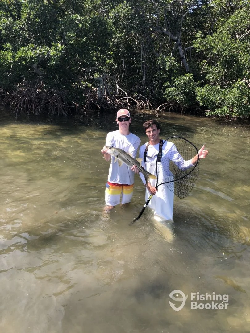 Two men standing in shallow water near mangrove trees, holding a fish and a fishing net, after a successful fishing trip.