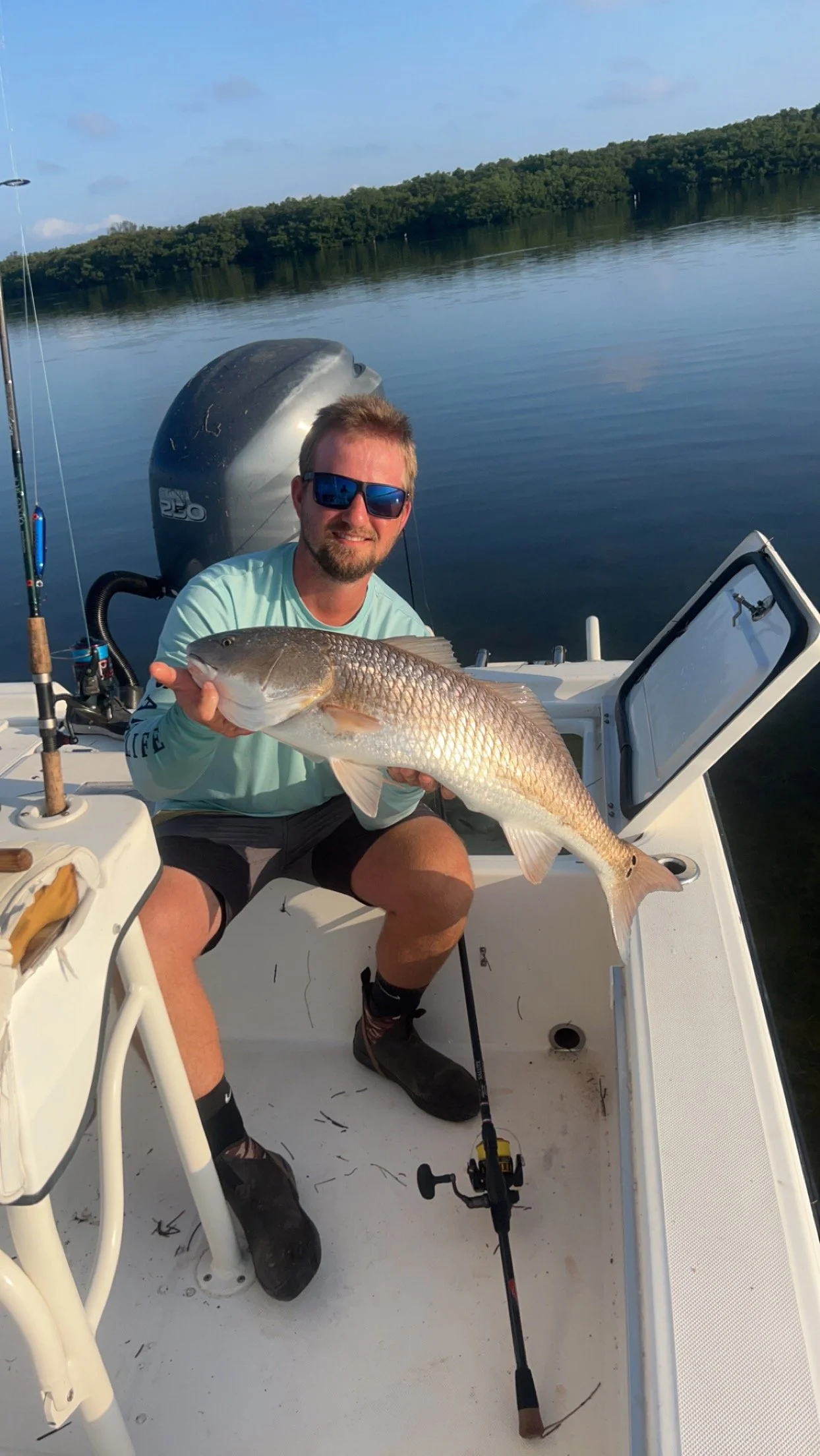 Man in sunglasses holding a large fish on a boat in a calm lake with trees in the background.