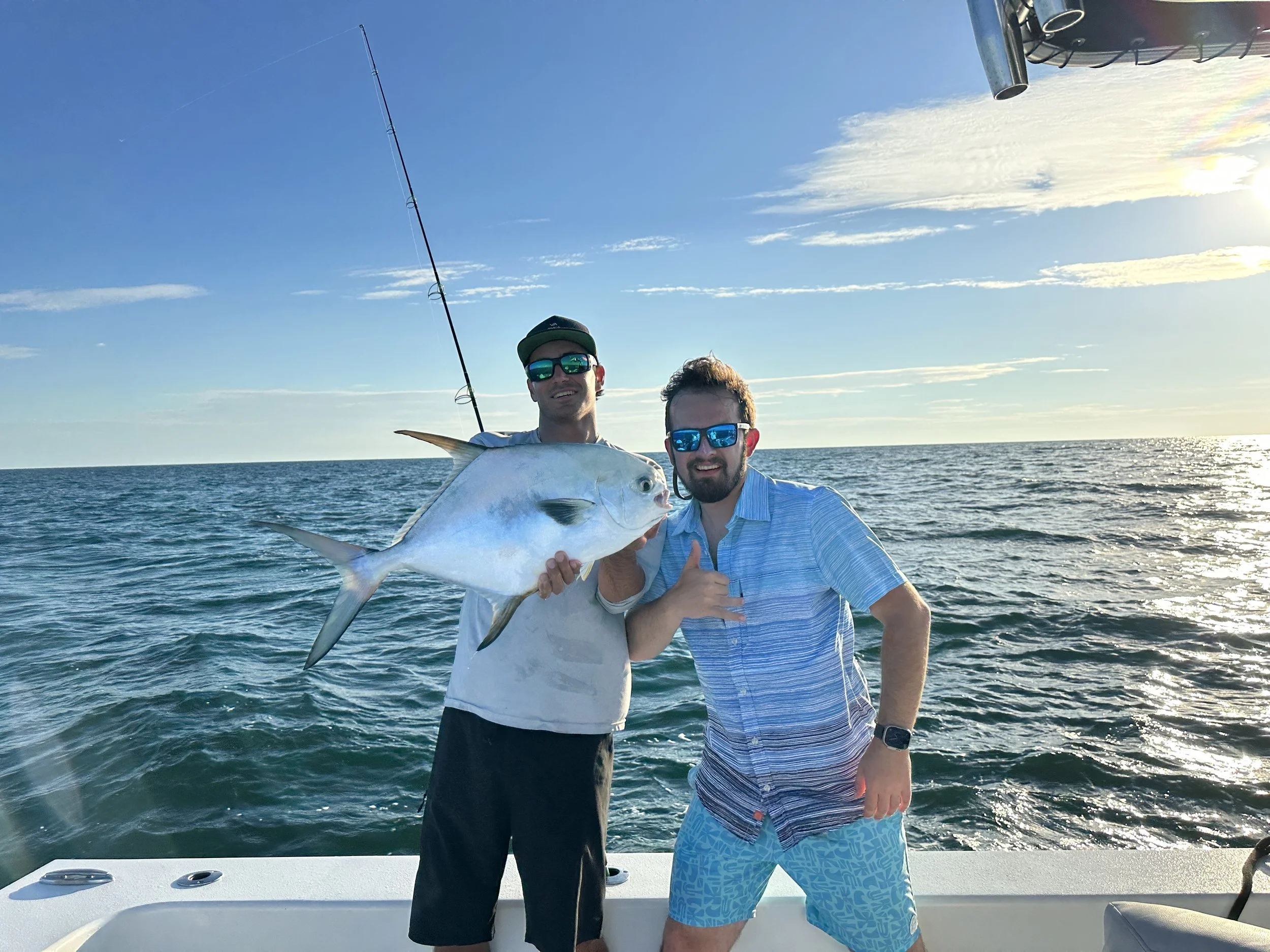 Two men on a boat at sea holding a large fish, with fishing rod in background, sunset sky, and calm water.