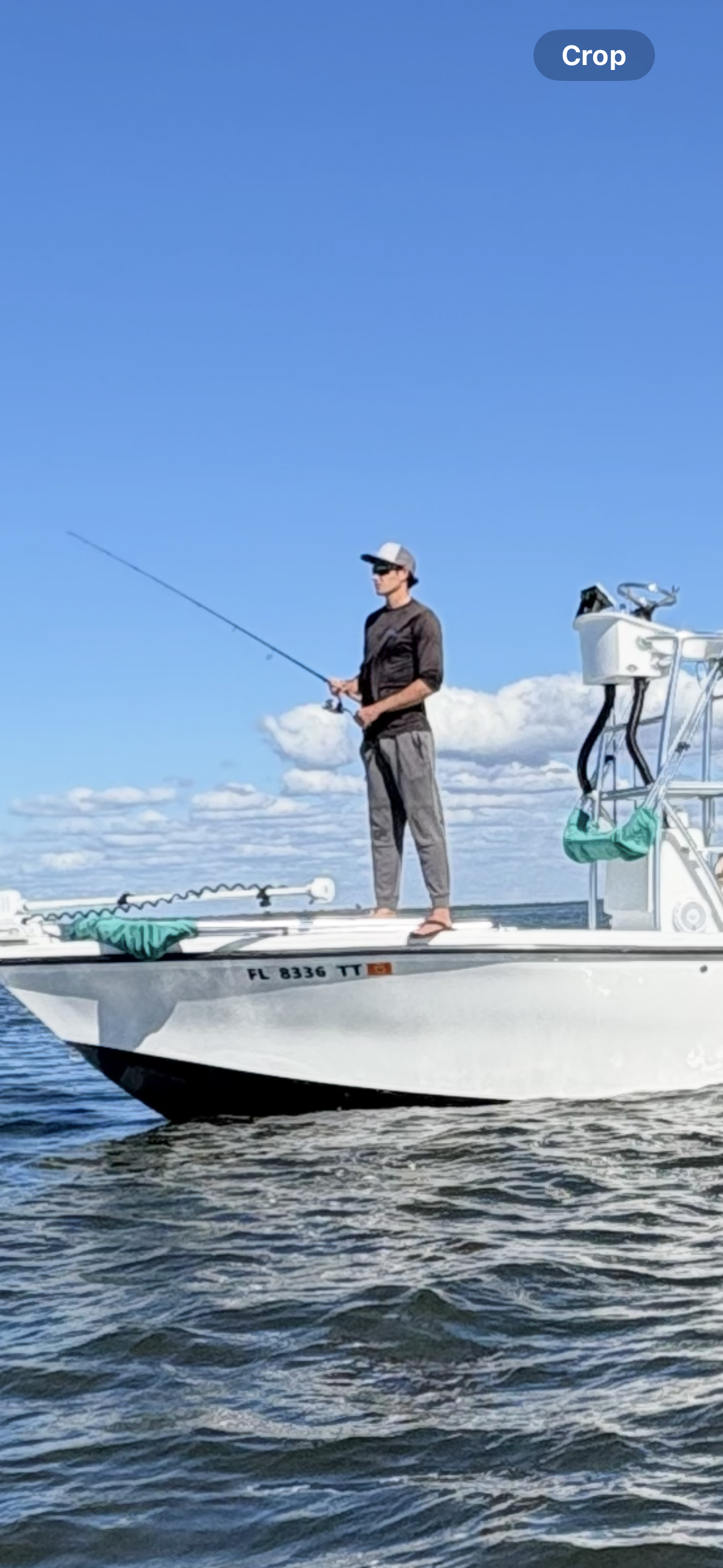 A person standing on a boat, fishing with a rod under a blue sky with clouds.