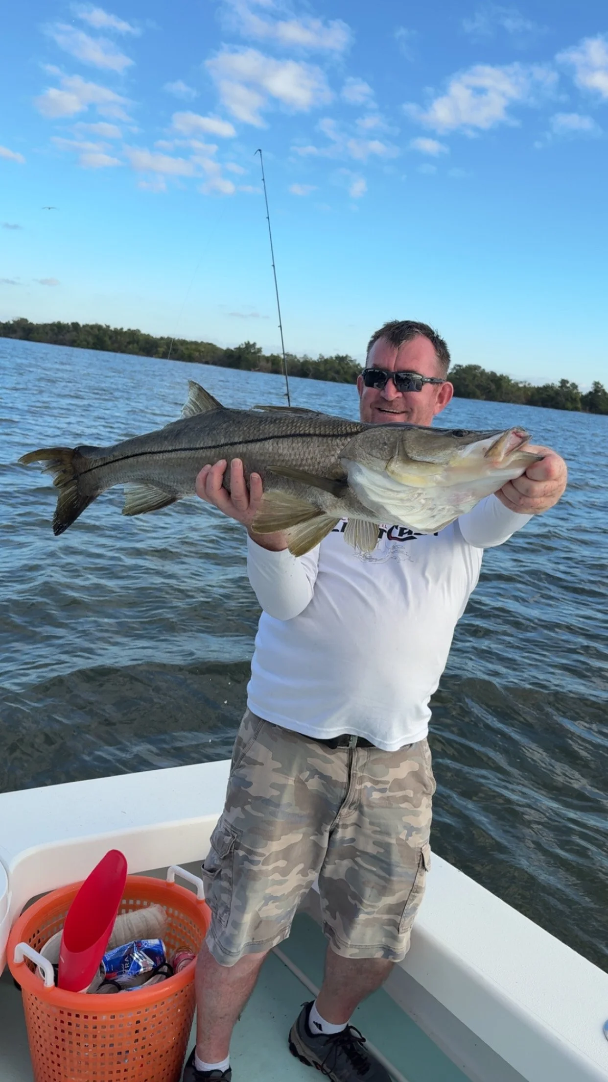 Man in sunglasses holding a large fish on a boat with water and trees in the background, a fishing pole is visible.