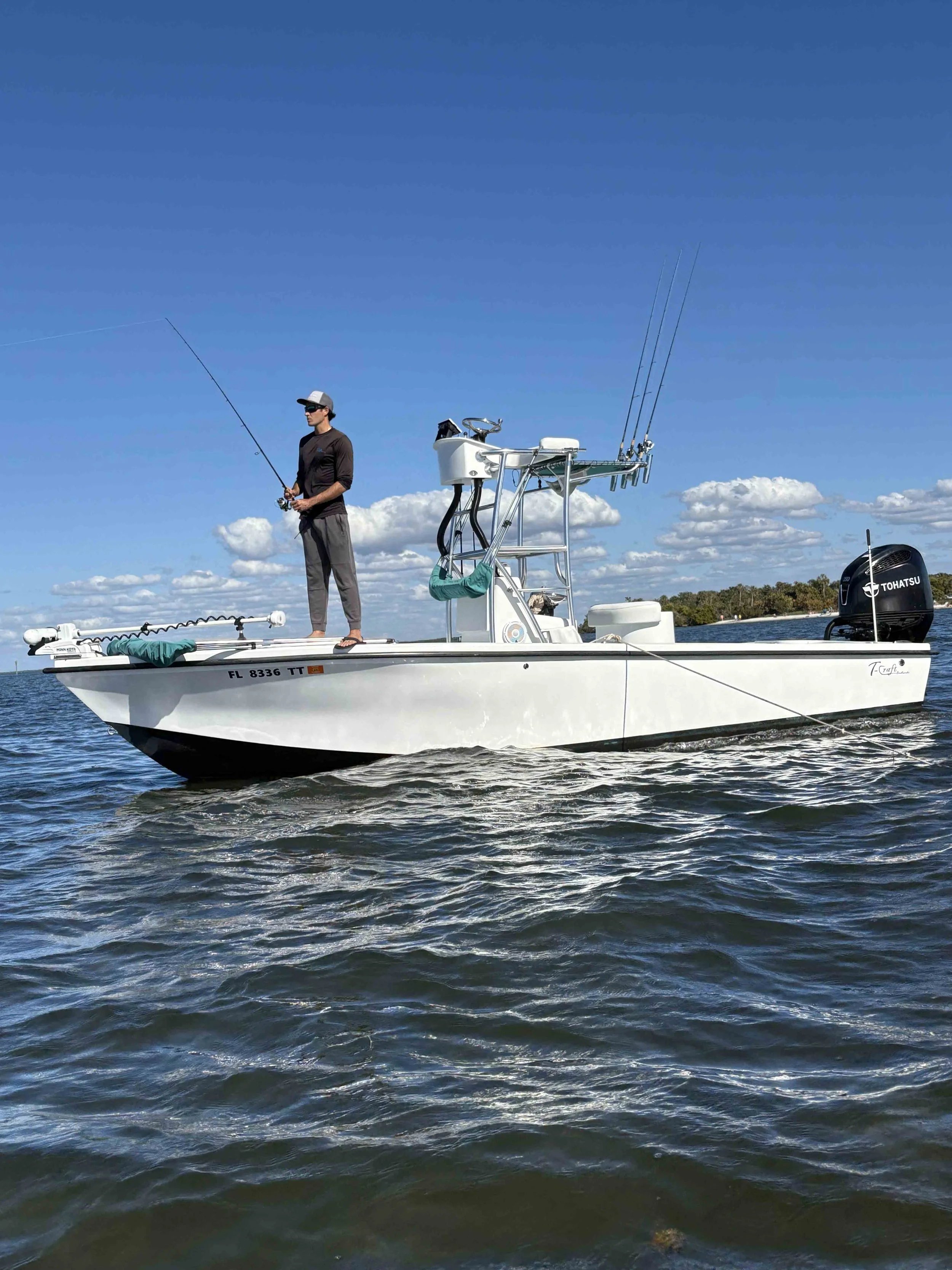 A person standing on a white fishing boat, fishing in the water under a blue sky with scattered clouds.