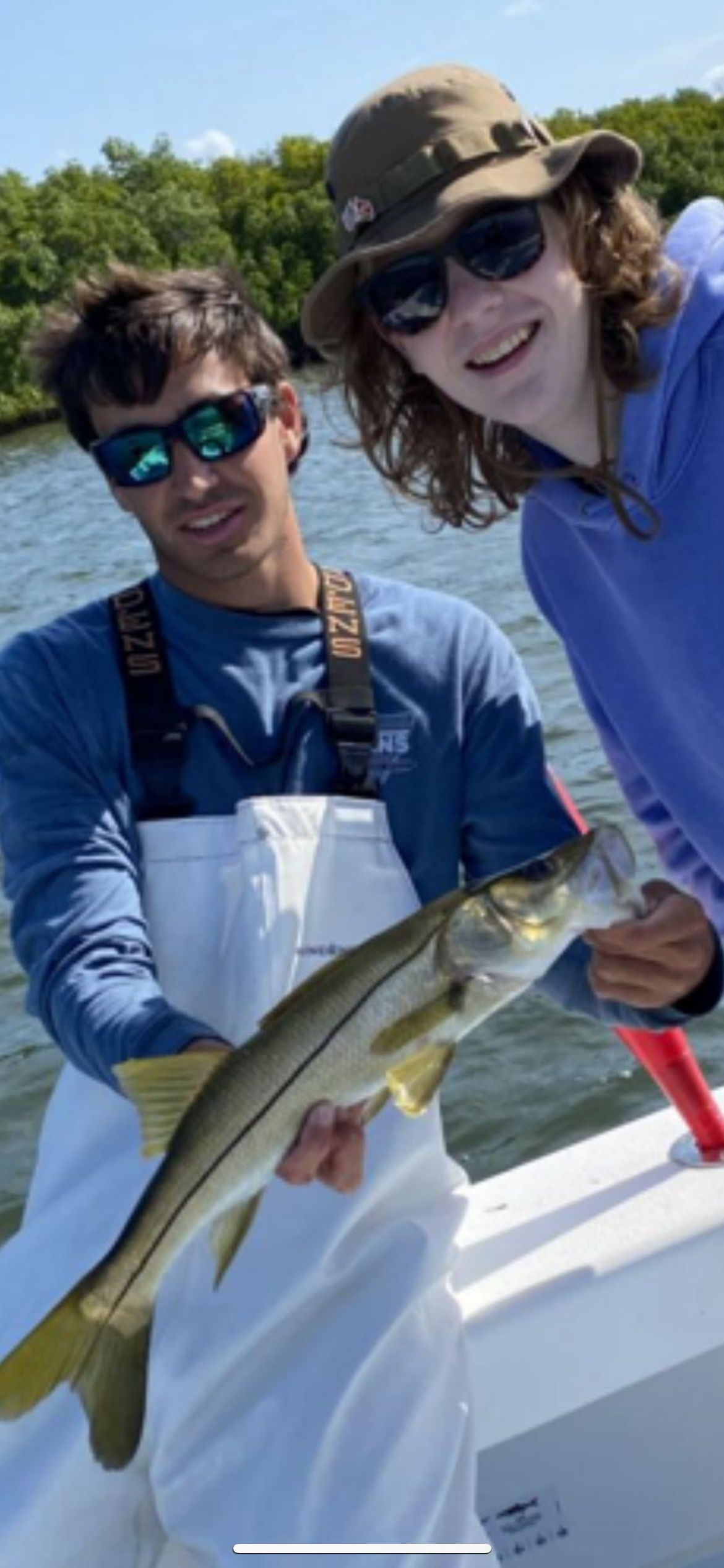 Two people on a boat, holding a fish they caught, with water and trees in the background. They are wearing sunglasses and outdoor clothing.