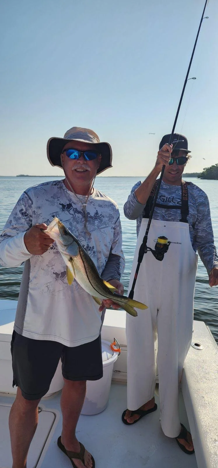 Two men on a boat holding a large fish, wearing fishing gear and sunglasses, with calm water and a clear sky in the background.