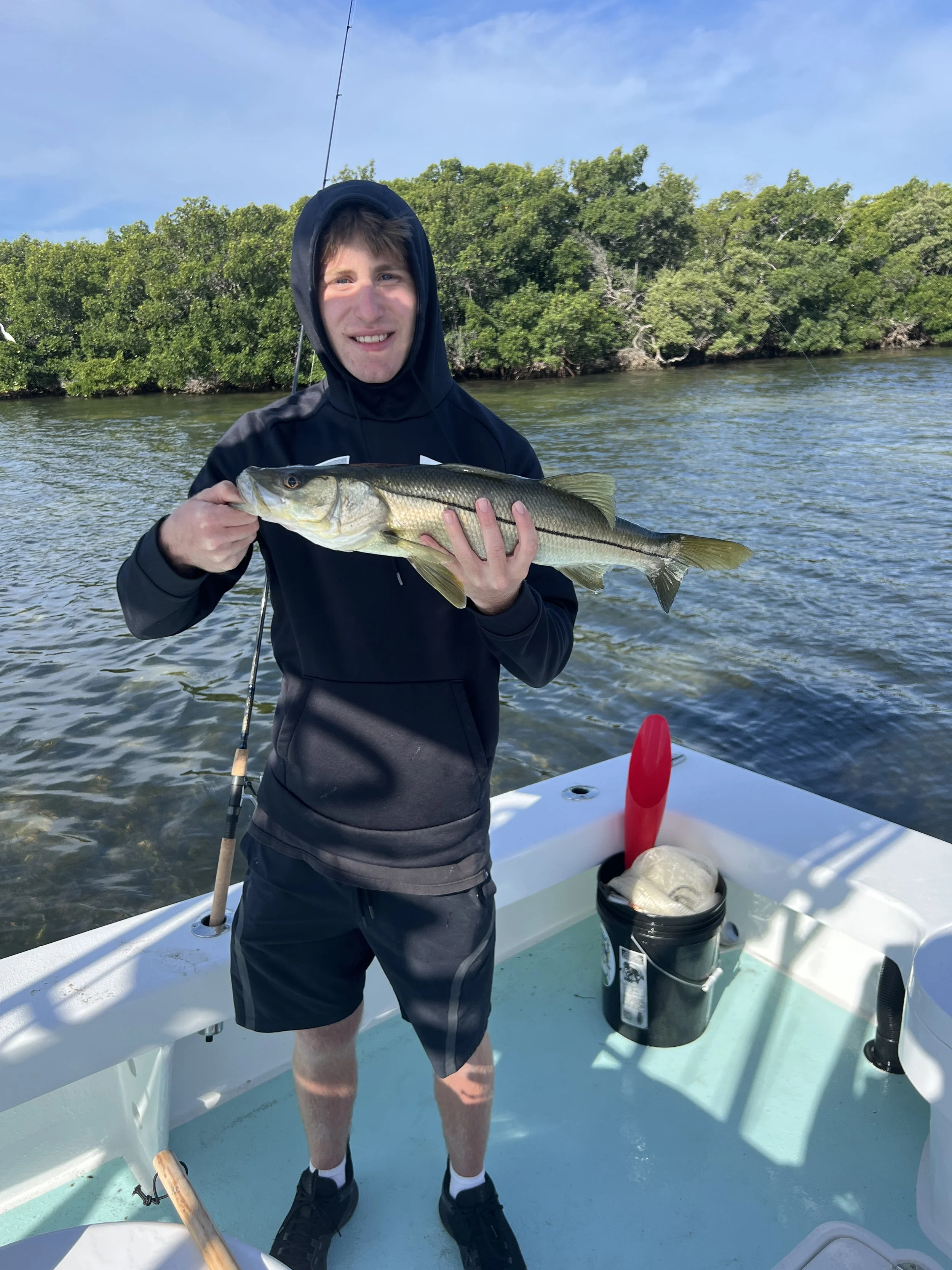 A young man in a black hoodie and shorts on a boat holding a large fish, with trees and water in the background on a clear day.