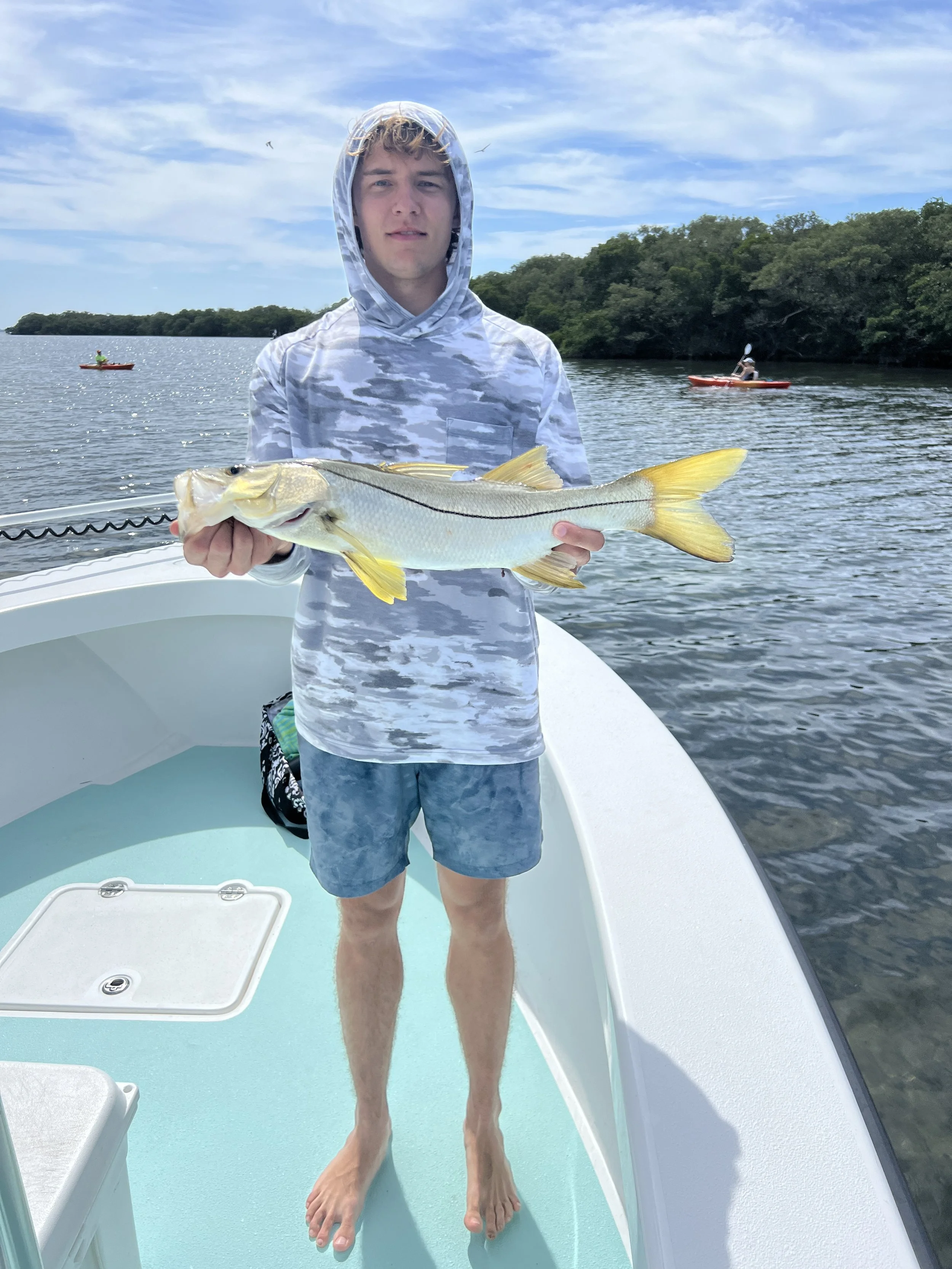 A young man standing on a boat holding a large fish with water and shoreline in the background.