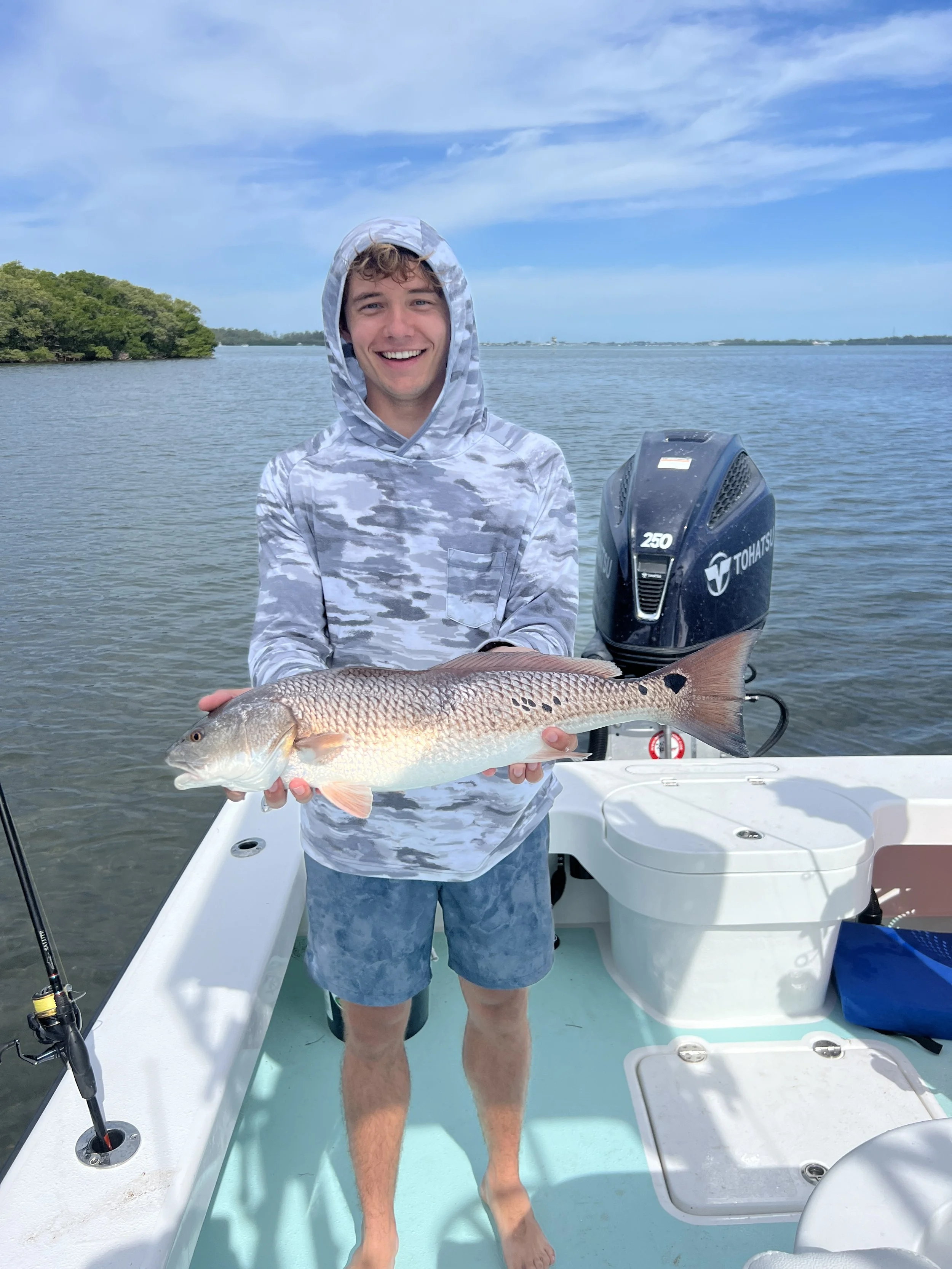 Young man holding a fish on a boat in a waterway with blue sky and some trees in the background.