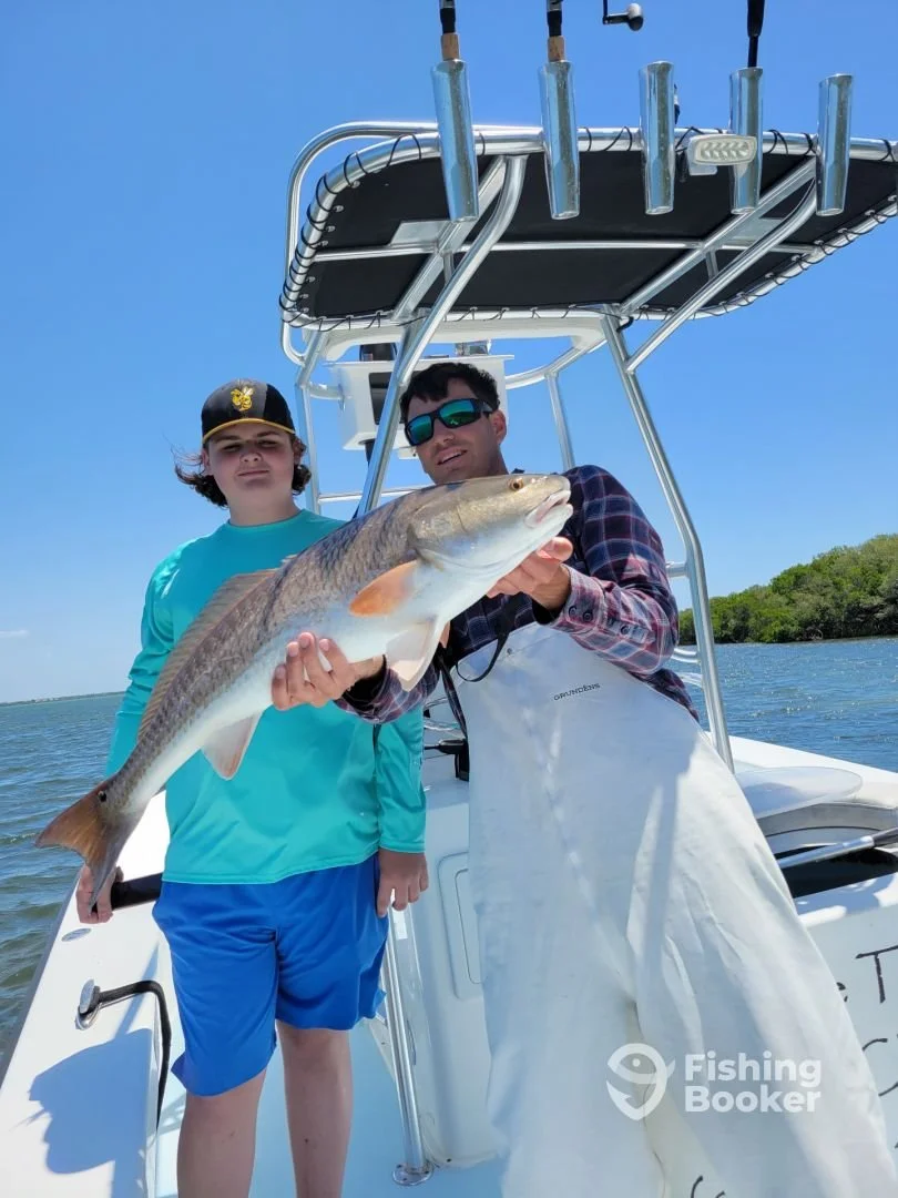 Two boys on a boat, one holding a large fish, with a blue sky and water in the background, during daytime.