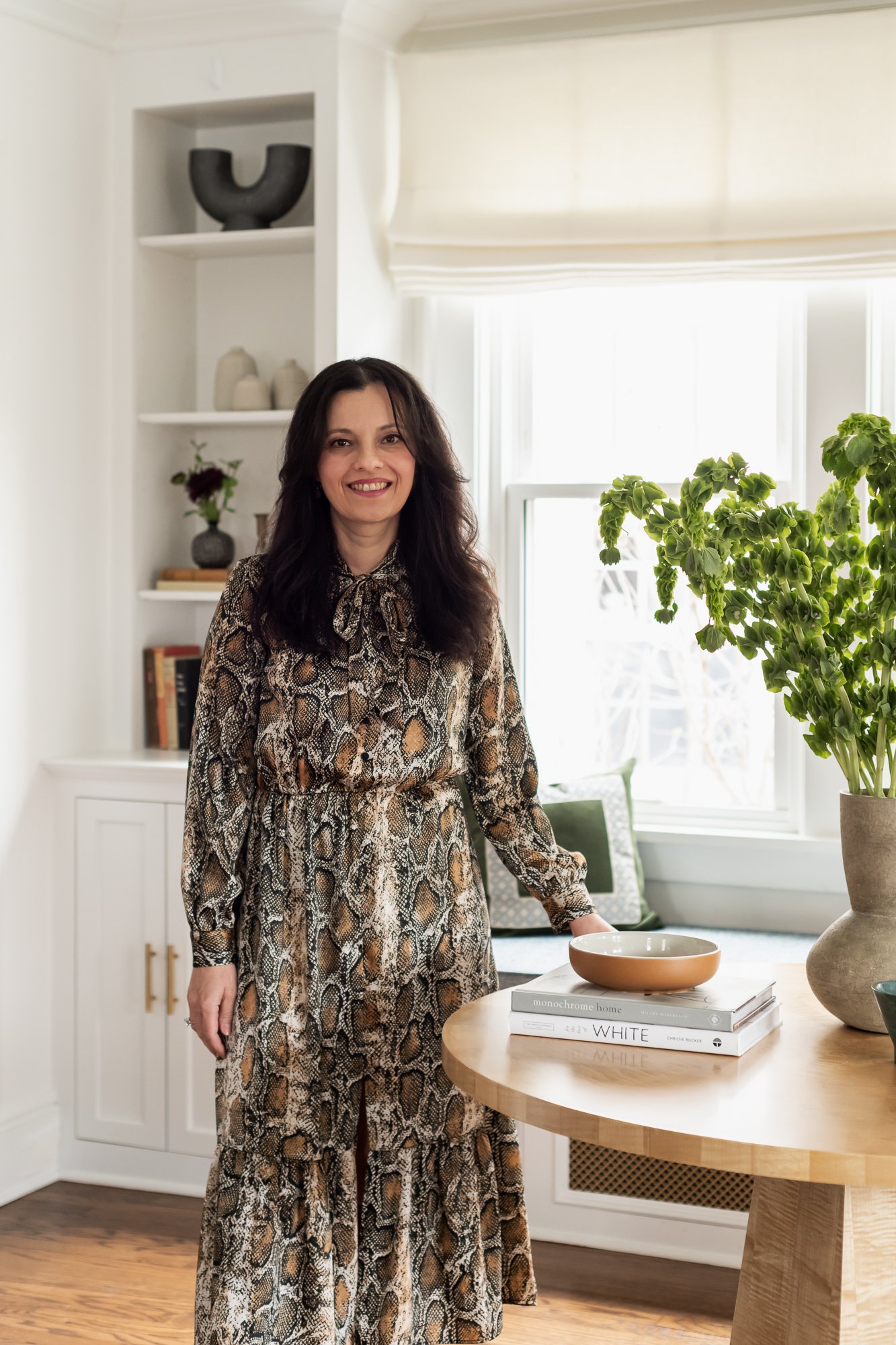 A woman with dark hair wearing a snake-print dress standing in a well-lit room beside a wooden table with books and a large potted plant.