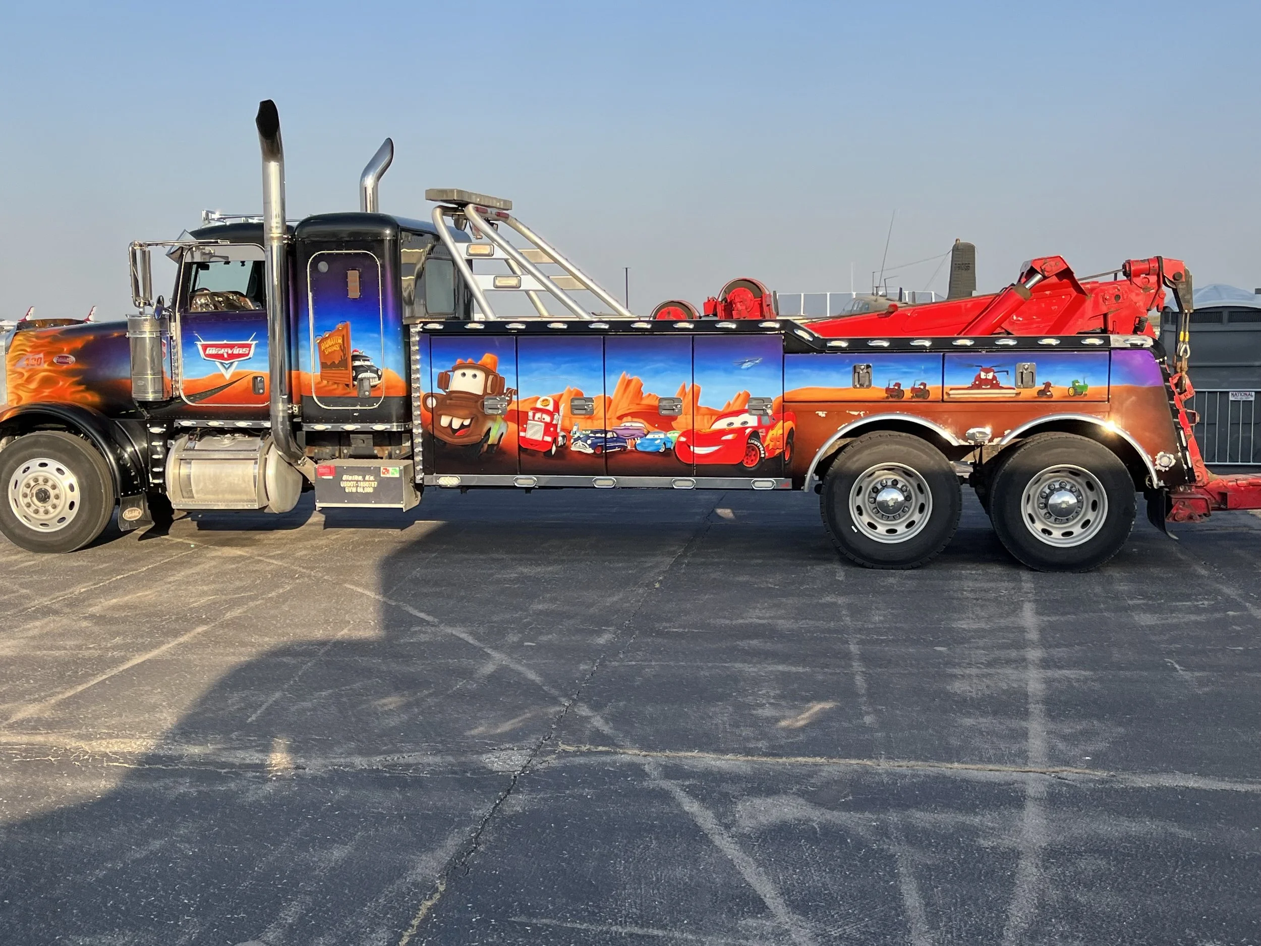 A tow truck decorated with characters from animated movies, featuring Lightning McQueen and Mater from Cars on the side, with a bright red boom and a background of outdoor sky.